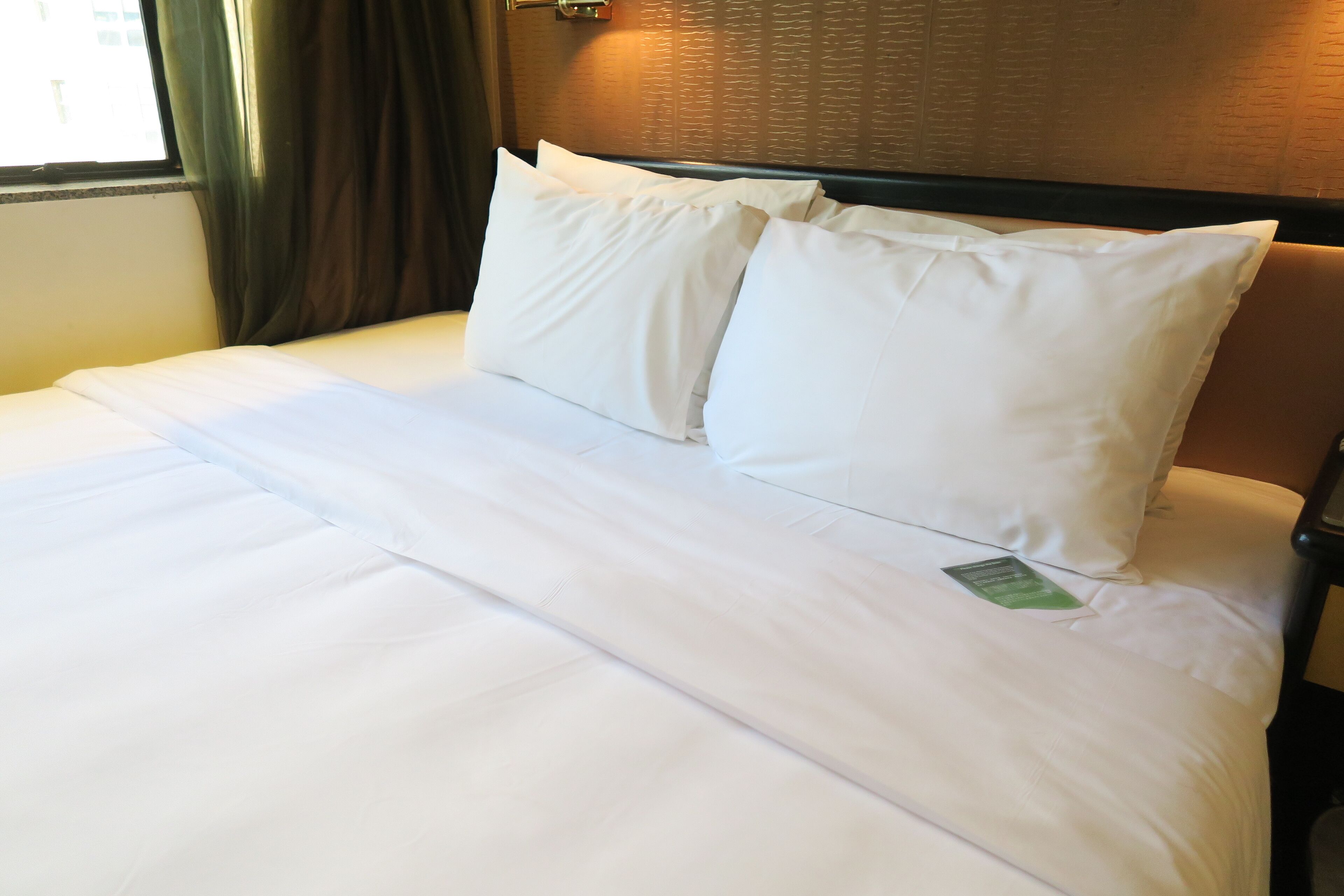 A neatly made bed with white linens and pillows, a wooden headboard, and a window with curtains.