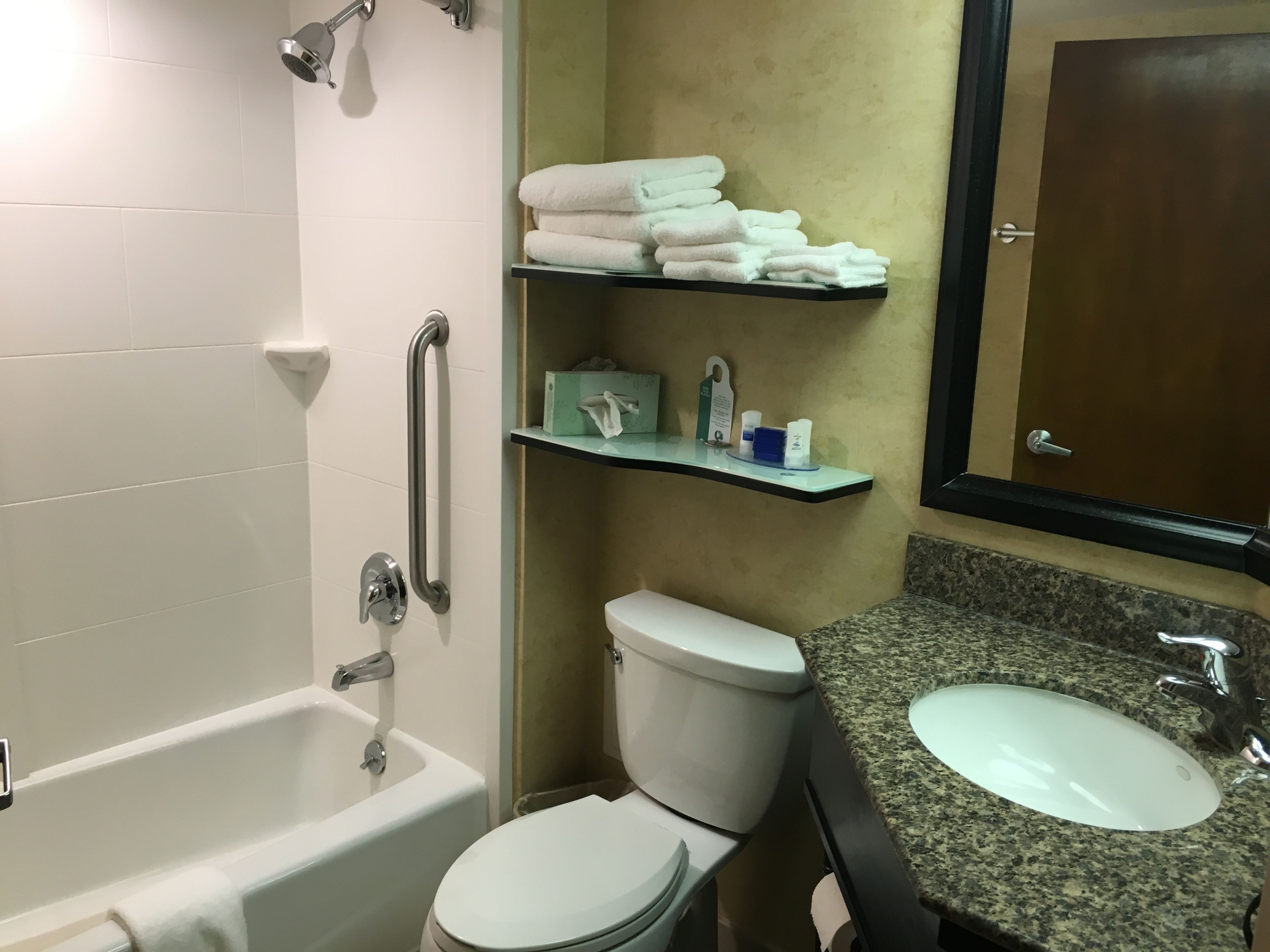 Un baño con bañera, inodoro y lavamanos. Hay repisas con toallas y artículos de aseo.