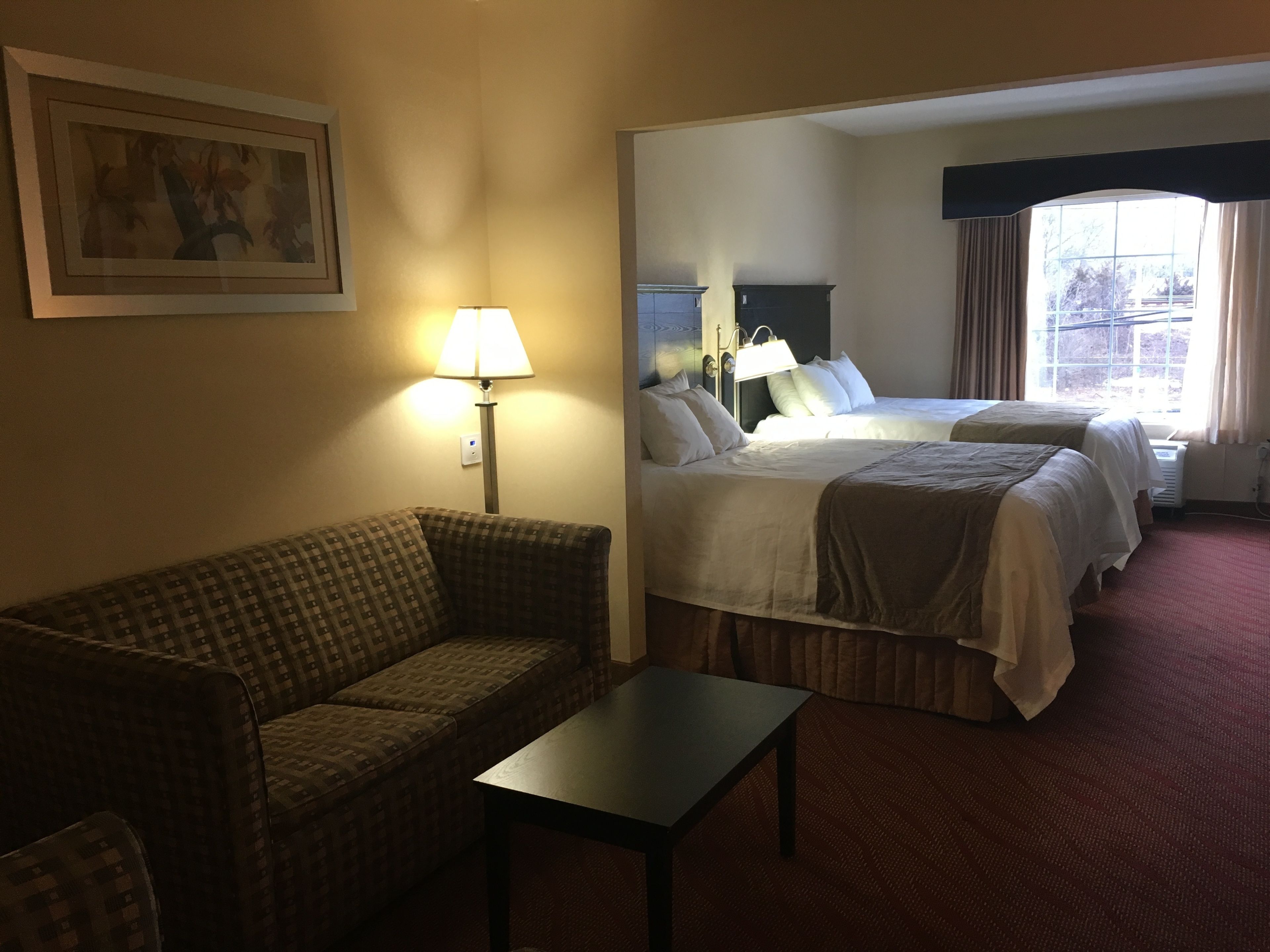 A hotel room with a sofa, a small table, two beds, a lamp, and a painting on the wall.