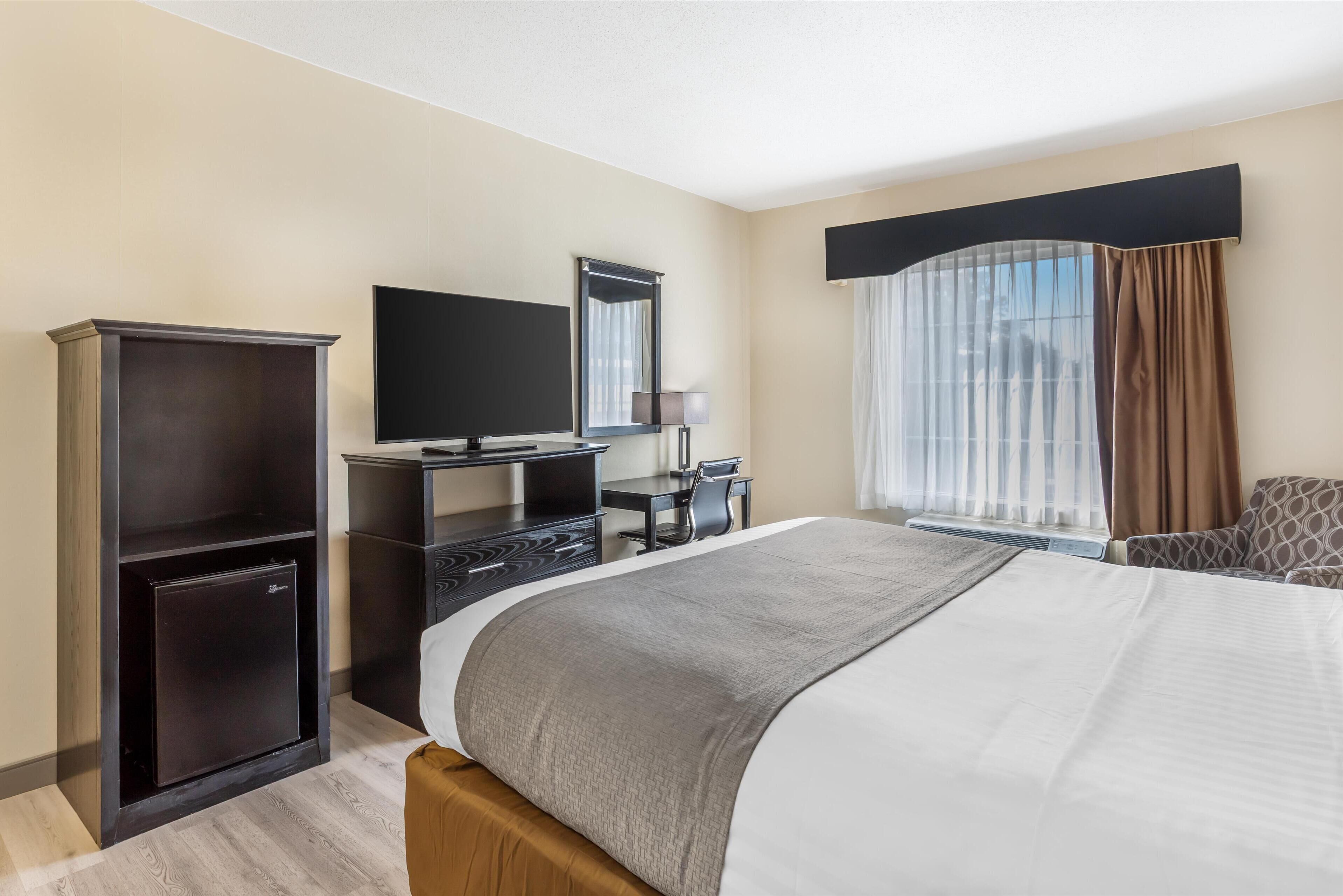 A hotel room with a large bed, a desk with a flat-screen TV, a small refrigerator, and a window with curtains.