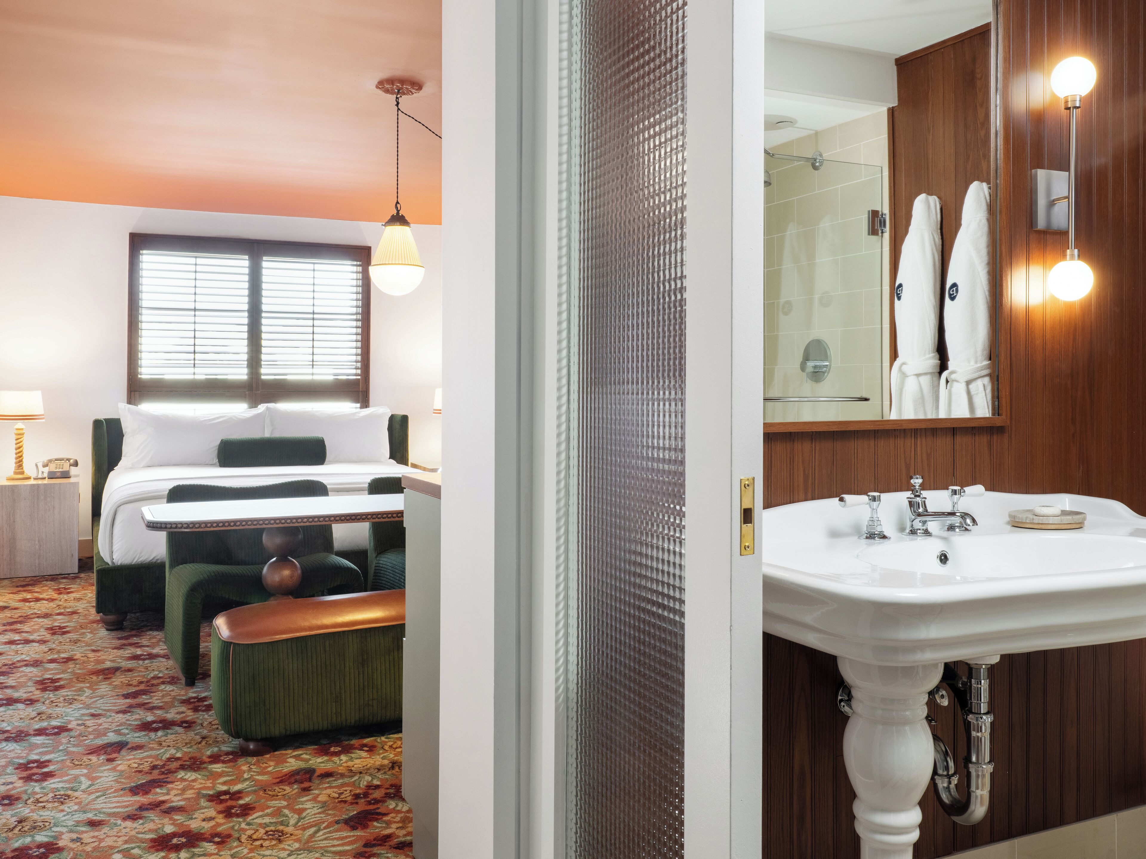 Una habitación de hotel con cama, una mesita, una silla, un banco, una ventana con persianas, una lámpara colgante, un baño con lavamanos, espejo y ducha.