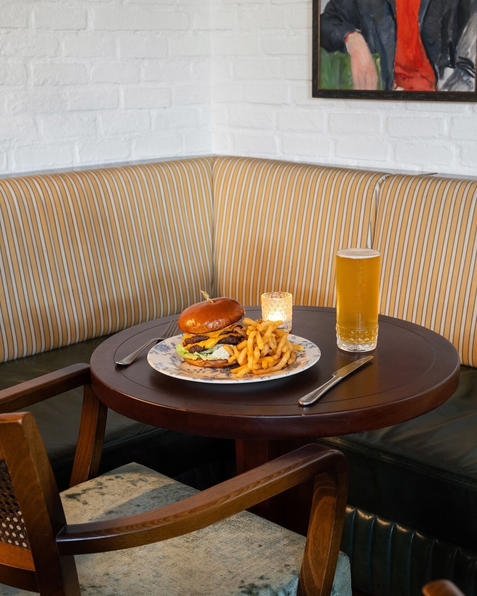 A corner seating area with a round table, a burger, fries, a glass of beer, and a candle.