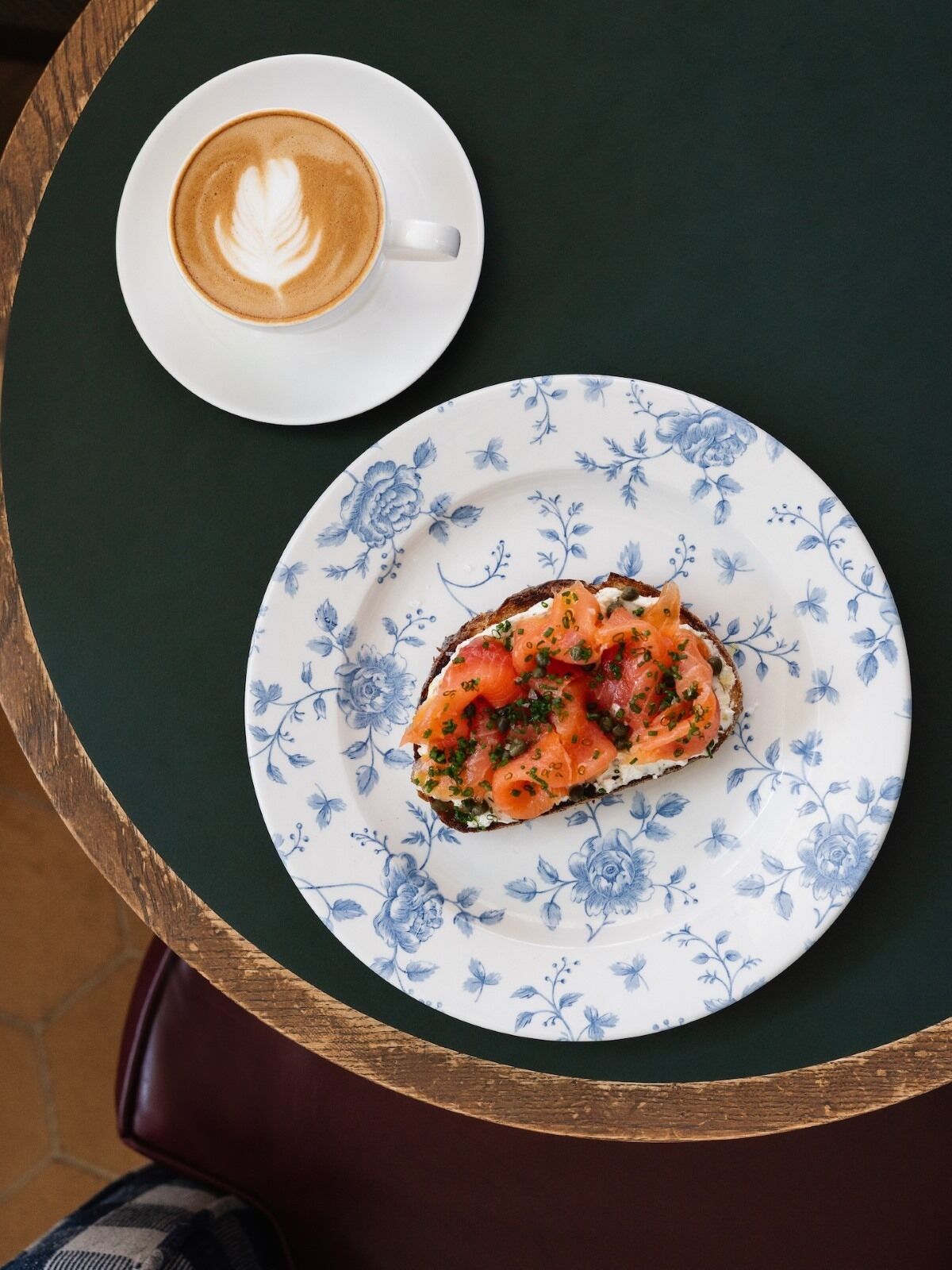 Una mesa con un plato de salmón ahumado sobre un plato estampado con motivos florales y una taza de café con arte latte.