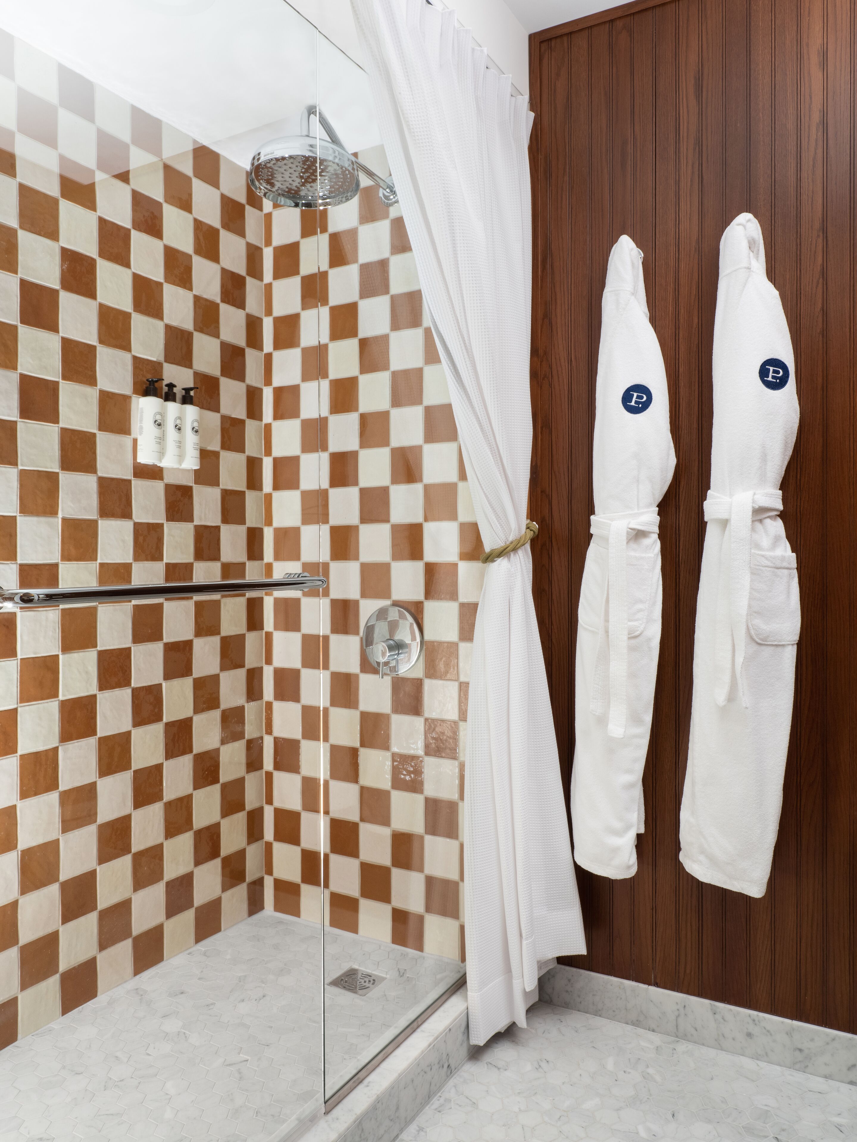 Zona de ducha con pared de baldosas a cuadros, puerta de cristal y puerta de madera. Hay dos toallas blancas colgadas en ganchos con un logo.