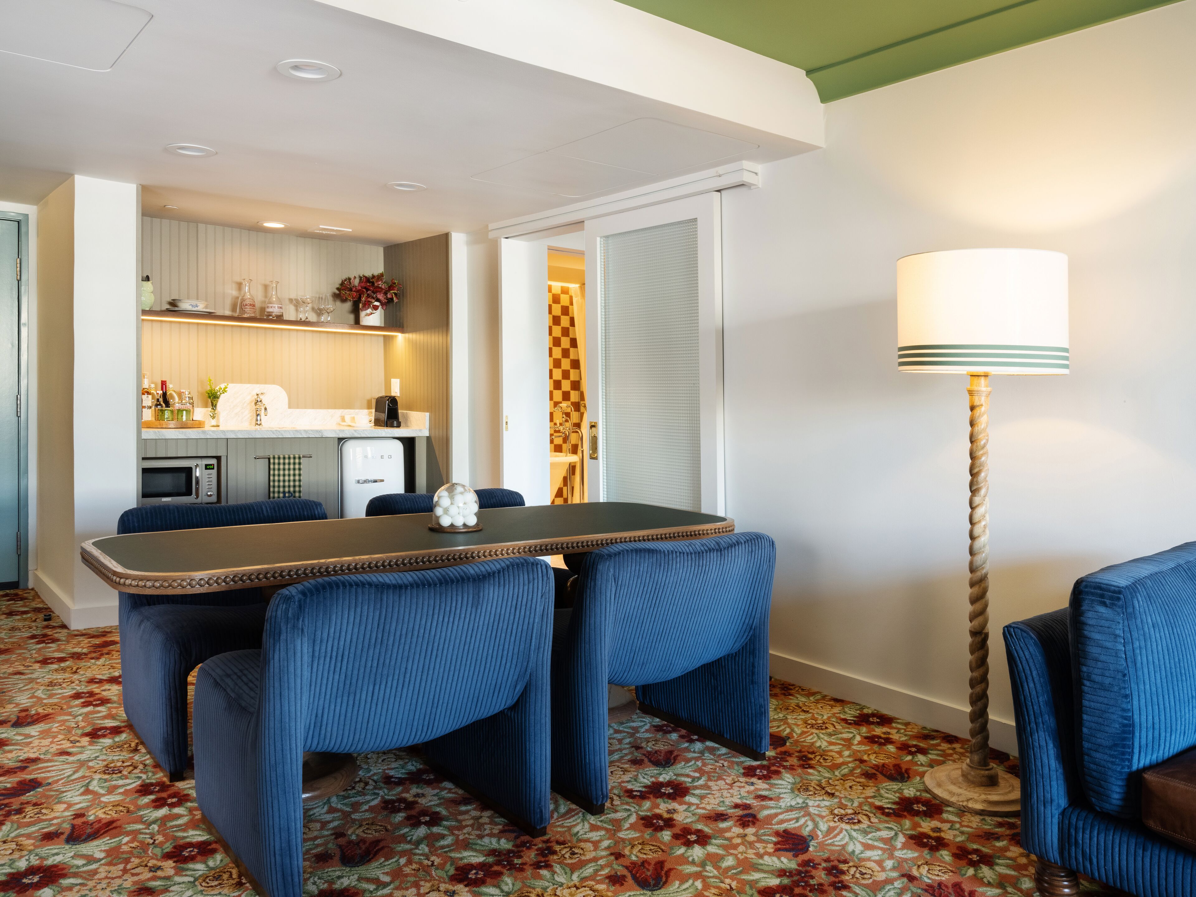 A hotel room with a dining area featuring a table and blue upholstered chairs, a kitchen area with a microwave and toaster, and a floor lamp.