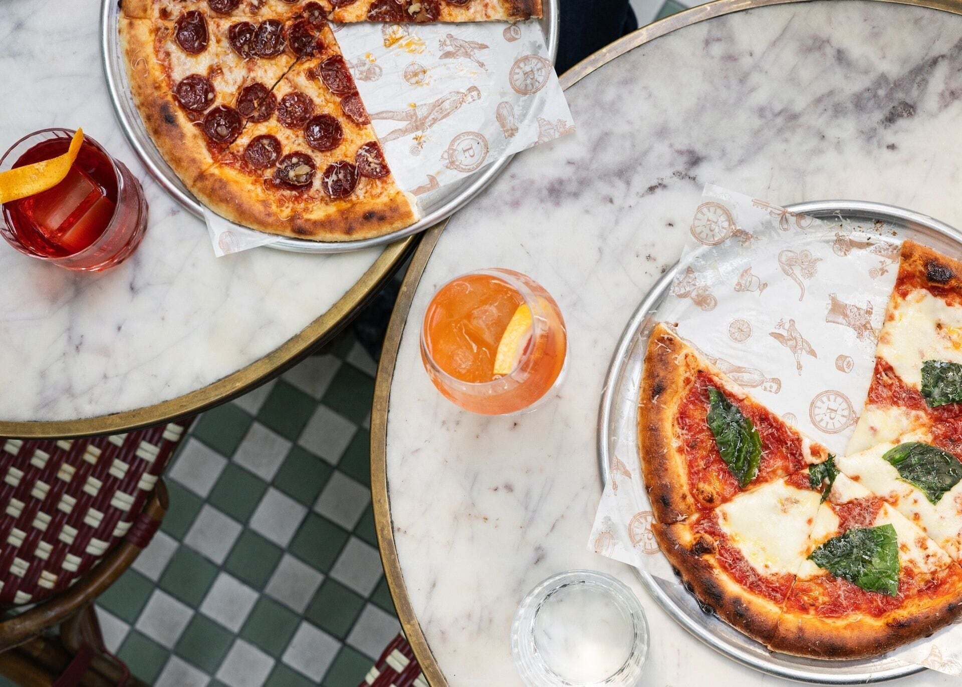 Eine Pizza auf einer Metallplatte, ein Getränk mit Orangenscheibe und ein Glas auf einem Marmortisch.