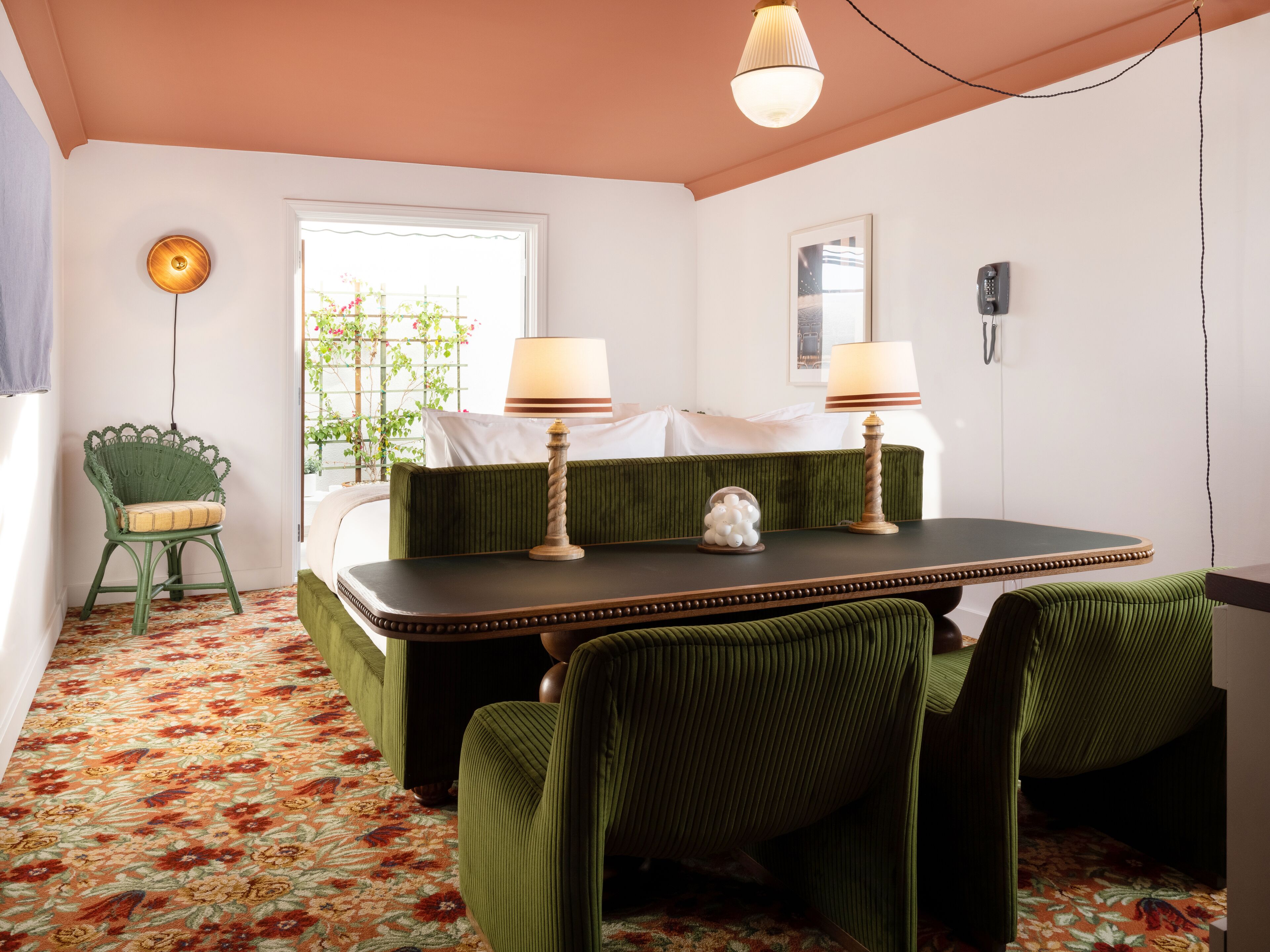 Une pièce avec un canapé recouvert d’un tissu vert, une petite table avec deux lampes, un tapis à motifs et une fenêtre donnant sur un paysage verdoyant.