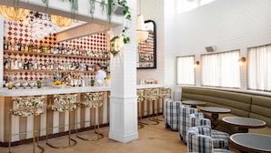 Un espace bar avec un mur en briques blanches, un barman et une variété de bouteilles et de verres exposés sur des étagères. On y trouve également des coins salon avec des chaises à motifs écossais et des tables rondes.