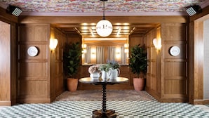 A hotel lobby with wooden paneling, a checkered floor, a central table with potted plants, and a floral ceiling.