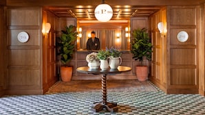 A hotel lobby with wooden paneling, a checkered floor, a central round table with potted plants, and a large chandelier.