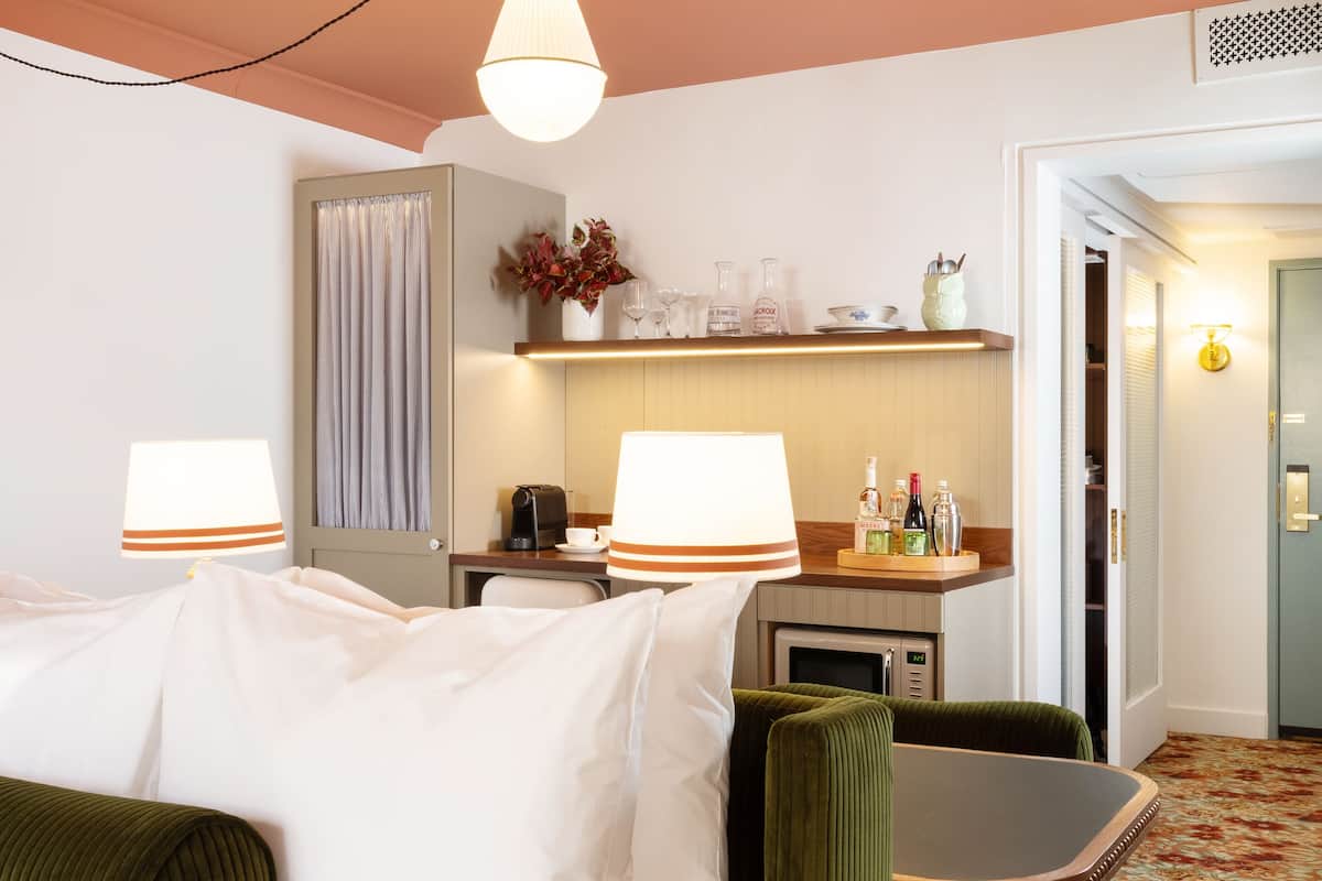 Een hotelkamer met een bed, een groene fauteuil, een klein tafeltje, een bureau, een lamp, een plank met decoratieve objecten en een deur naar een andere kamer.