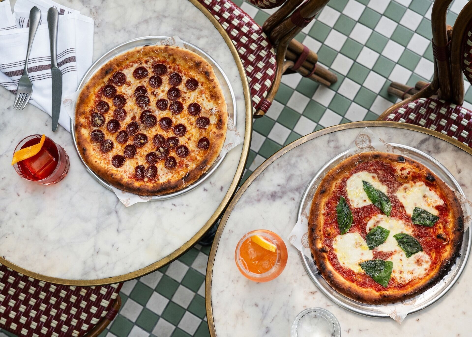 Dos pizzas sobre bandejas metálicas redondas en una mesa de mármol con mantel a cuadros.