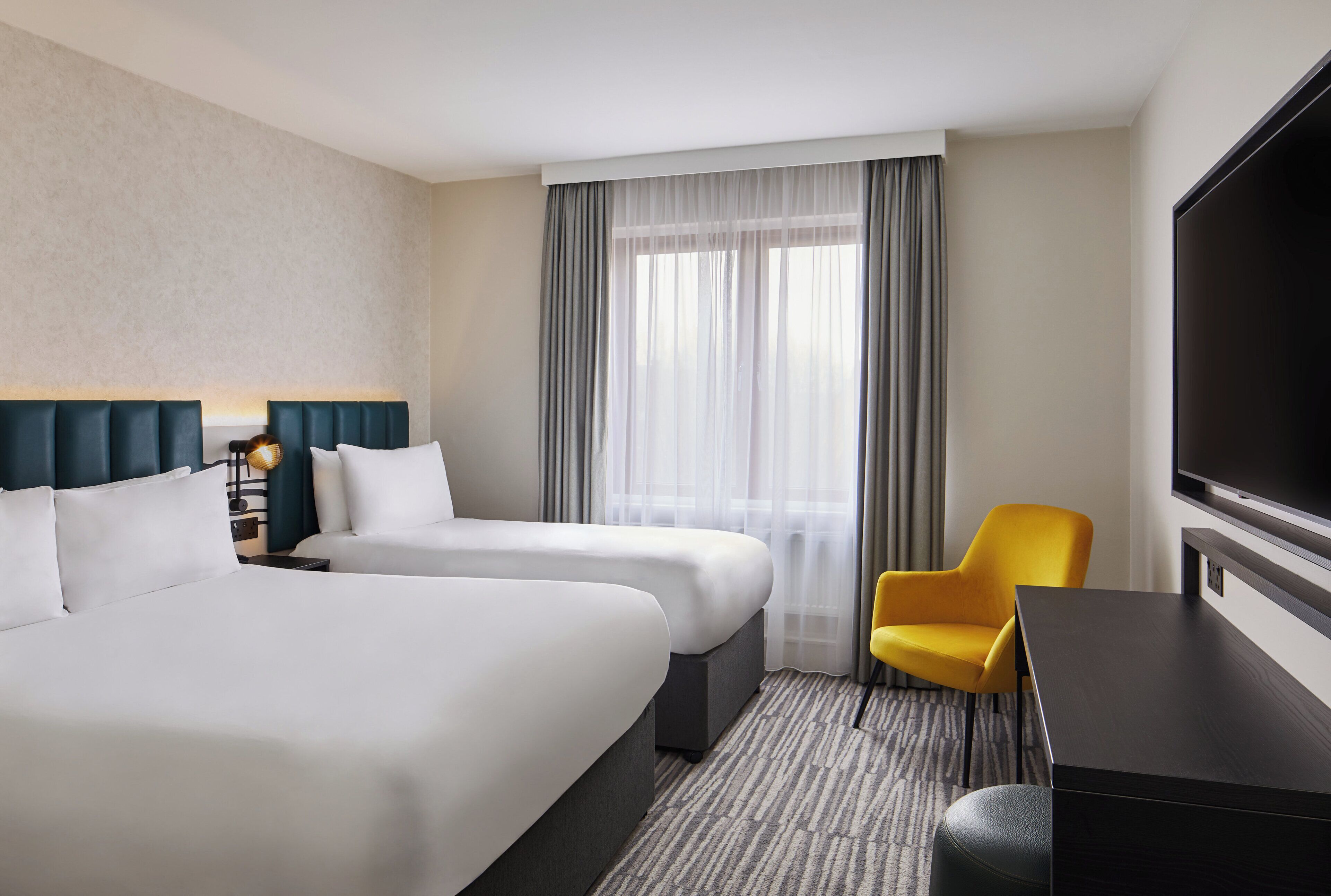 A hotel room with two beds, a desk, a chair, a TV, and a large window with curtains.
