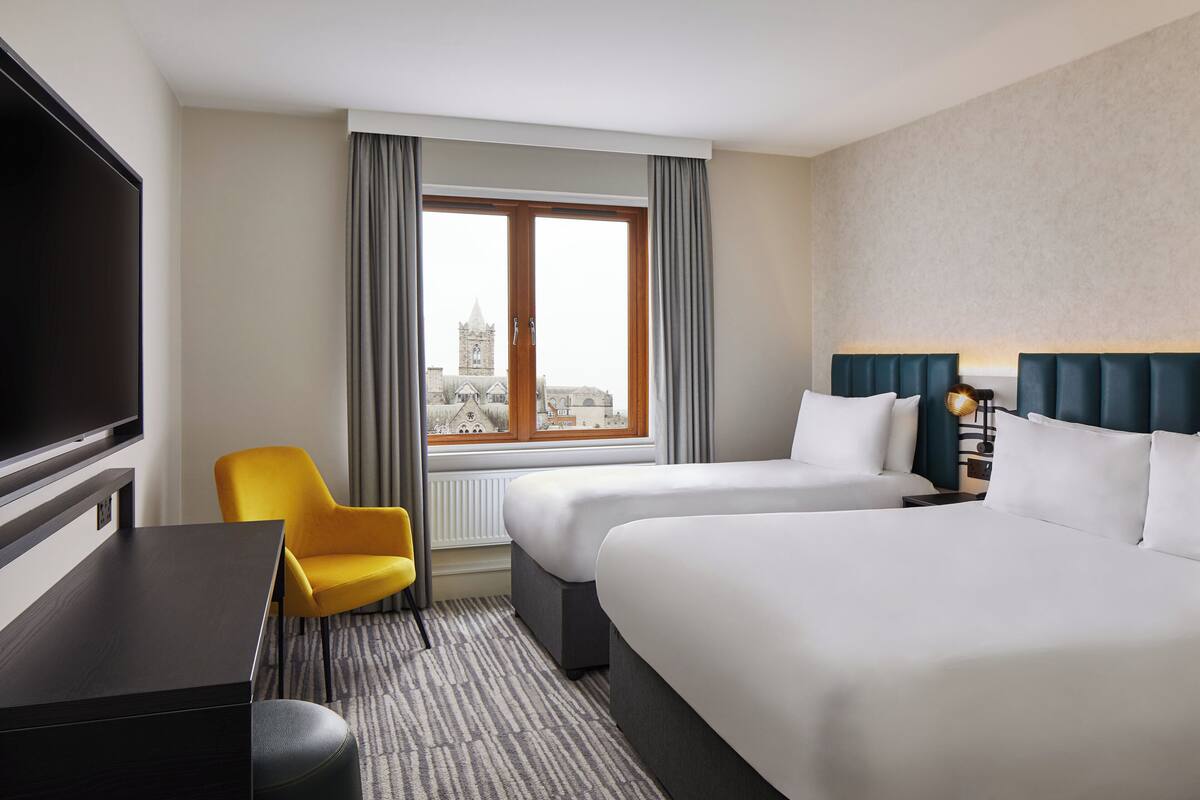 A hotel room with two beds, a TV, a desk, a chair, and a window with a view of a building.