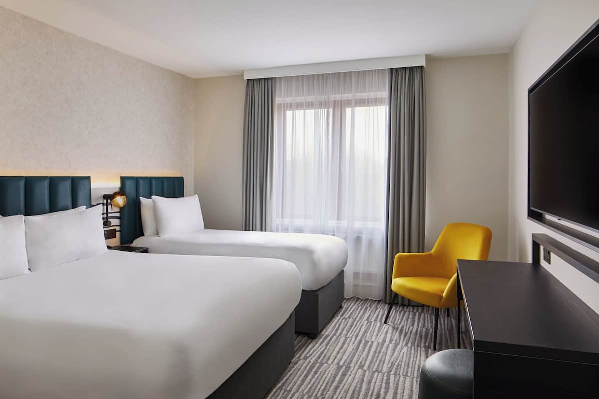 A hotel room with two beds, a desk, a chair, a TV, and a large window with curtains.