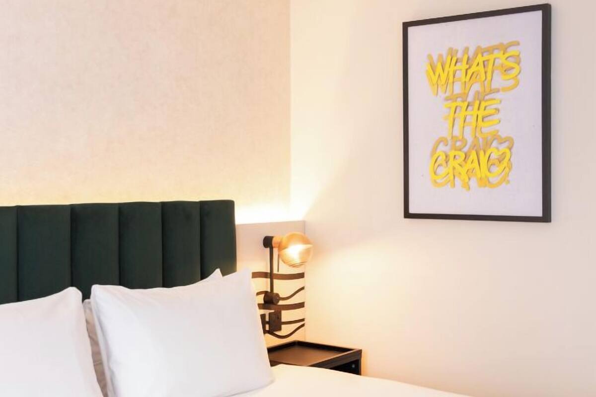 A hotel room with a bed, a headboard, a wall-mounted light, and a framed quote on the wall.