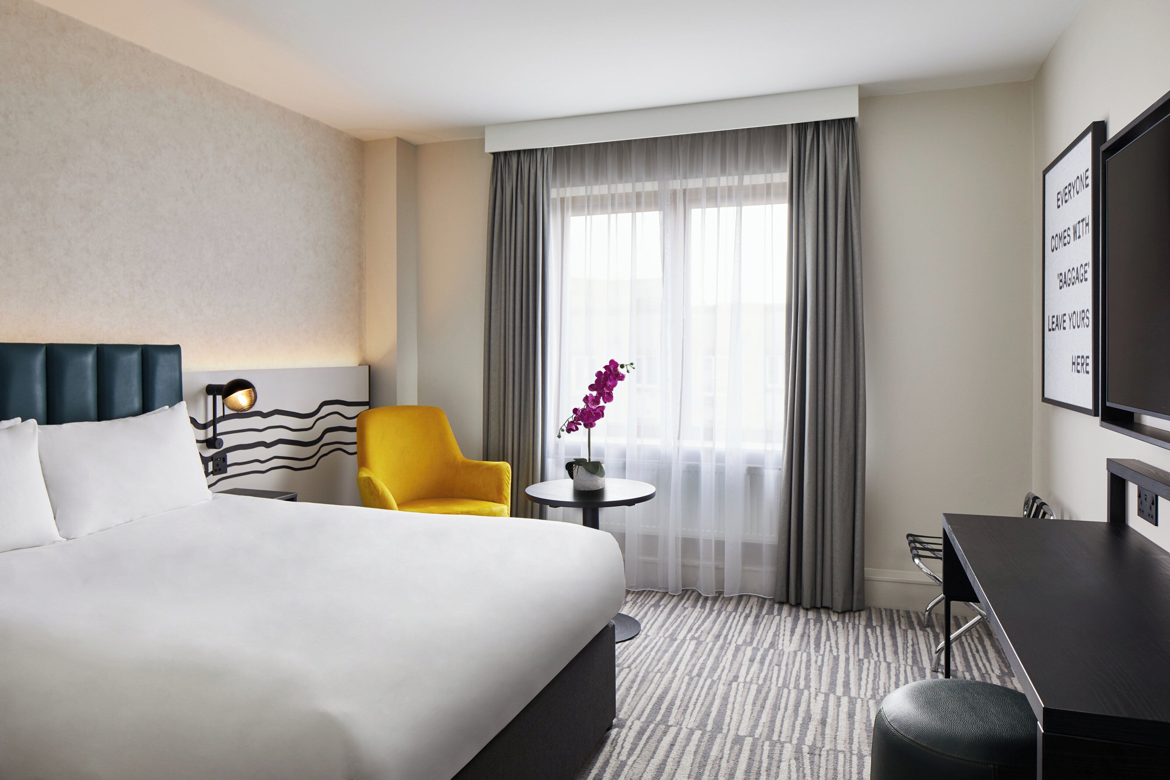 A hotel room with a large bed, a yellow armchair, a small table with a vase, a desk, and a TV on the wall.