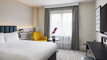 A hotel room with a large bed, a yellow armchair, a small table with a vase, a desk, and a TV on the wall.
