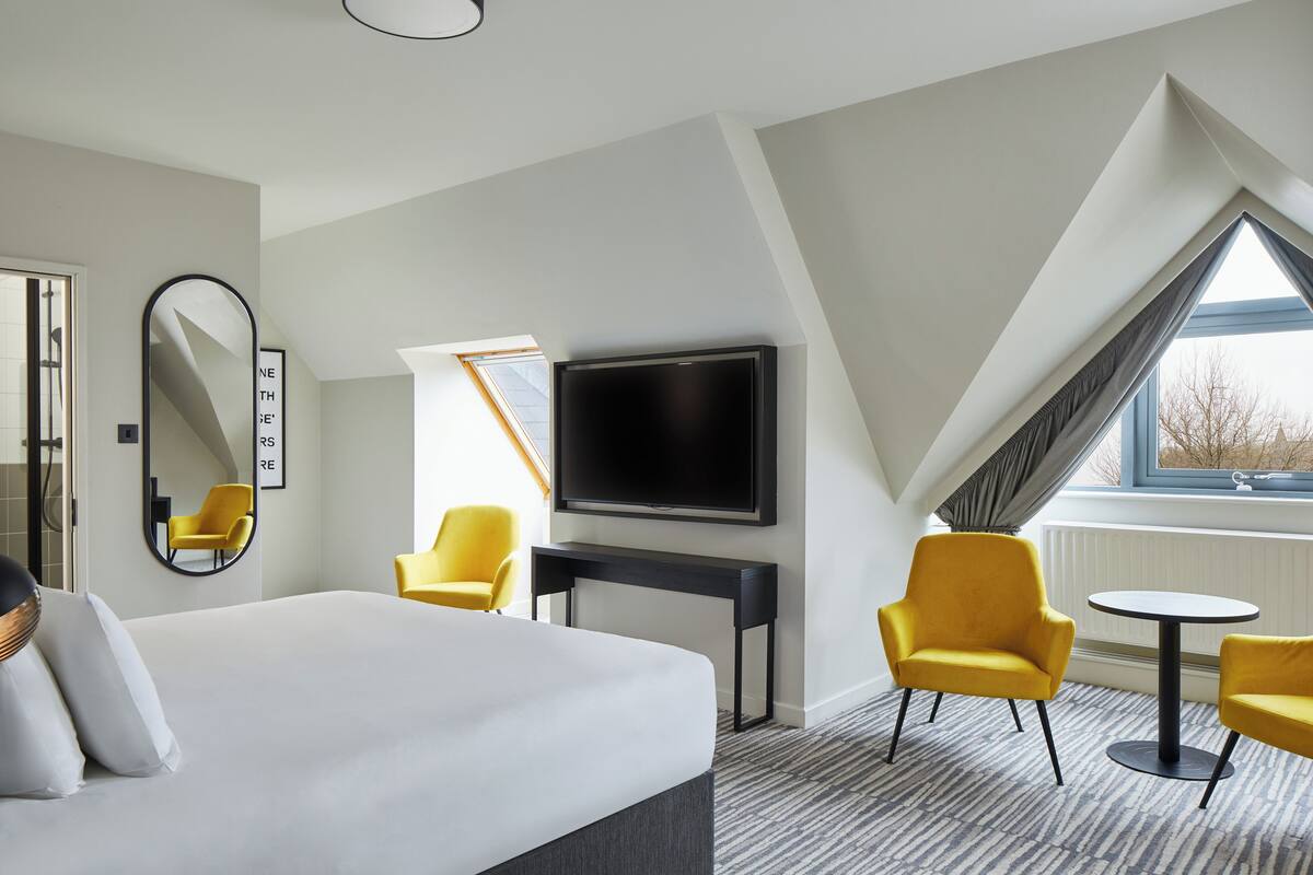 A hotel room with a large bed, a flat-screen TV mounted on the wall, two yellow armchairs, a small round table, and a large window with a view of the outside.