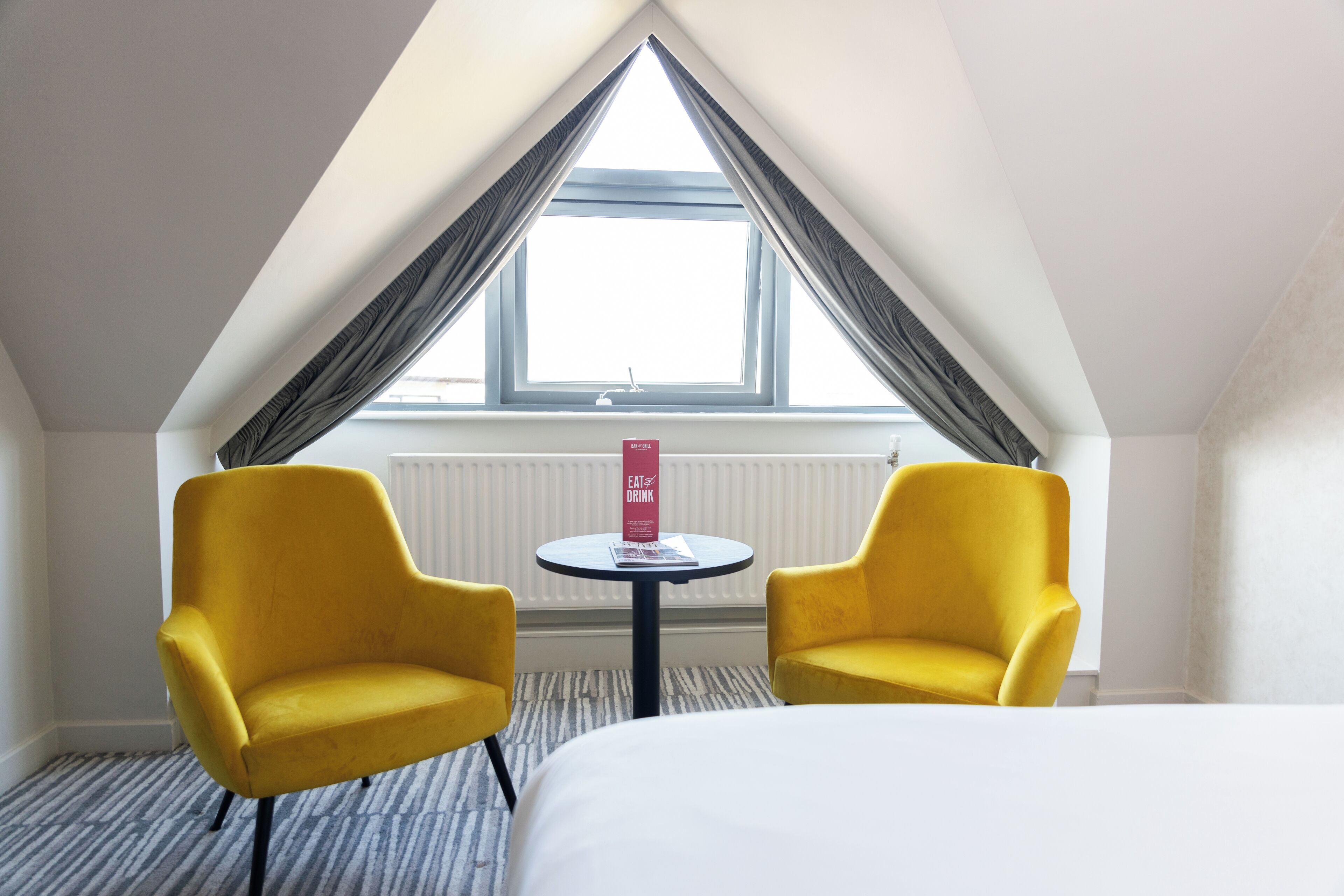 A hotel room with a bed, two yellow armchairs, a small table with a sign, and a large window with curtains.