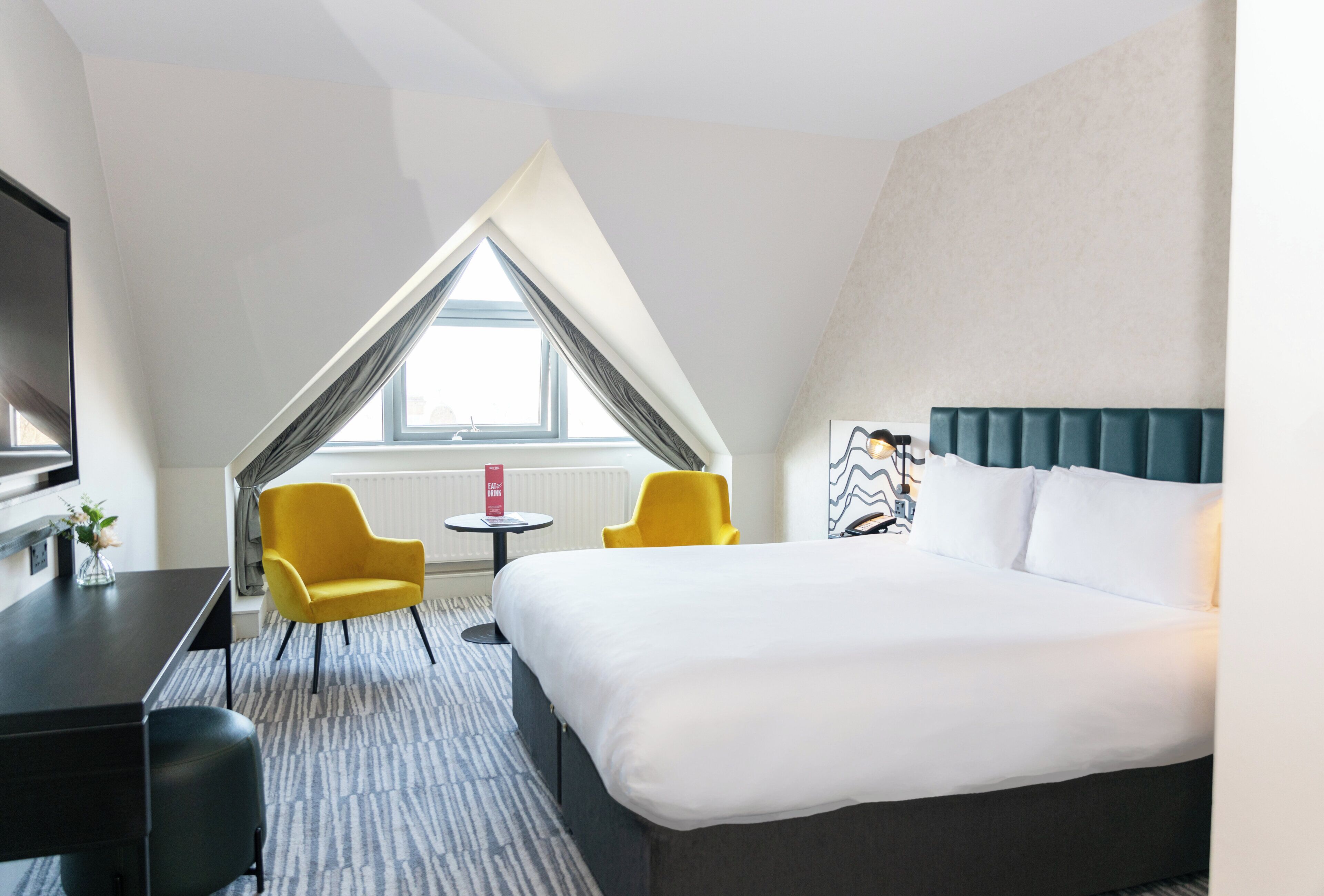 A hotel room with a large bed, two yellow chairs, a small table, and a TV.