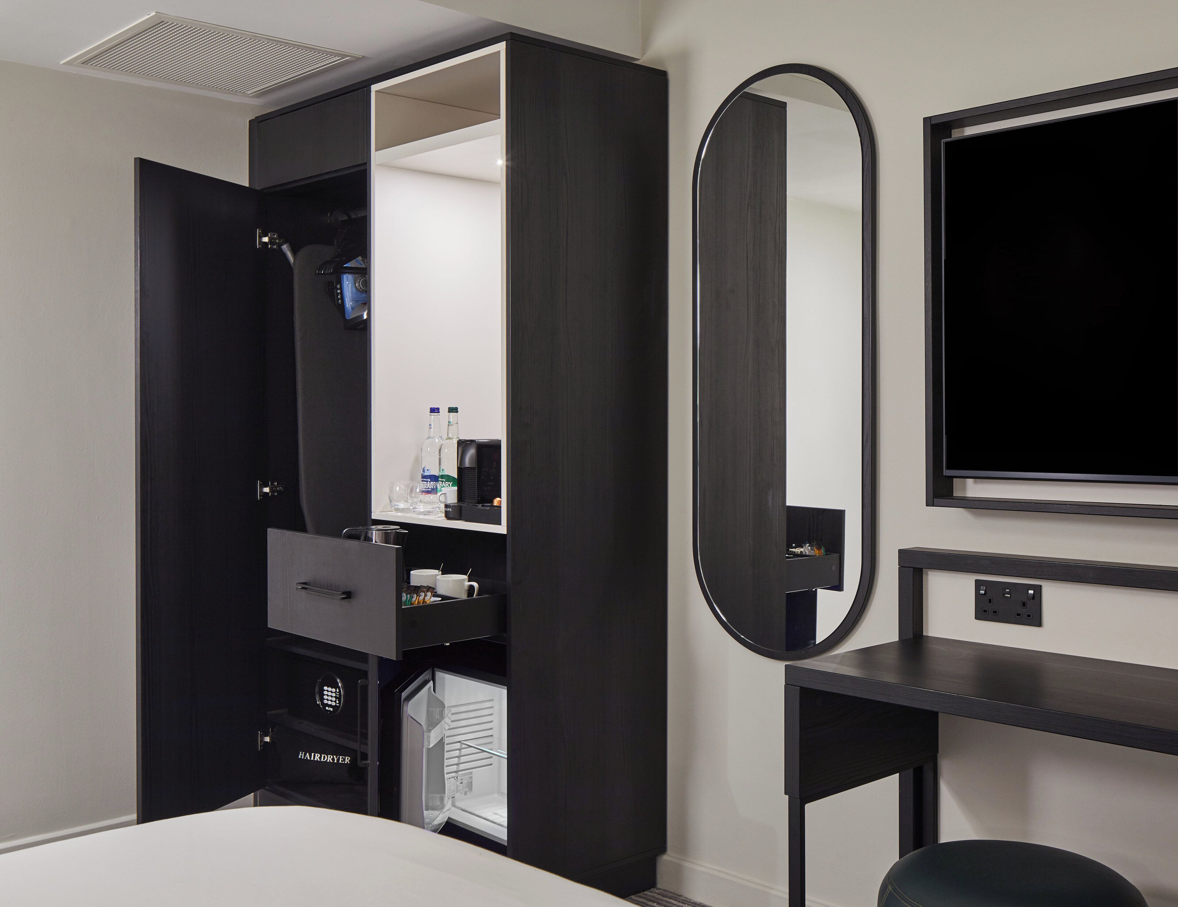 A hotel room with a built-in wardrobe, a mirror, a television, and a hairdryer.