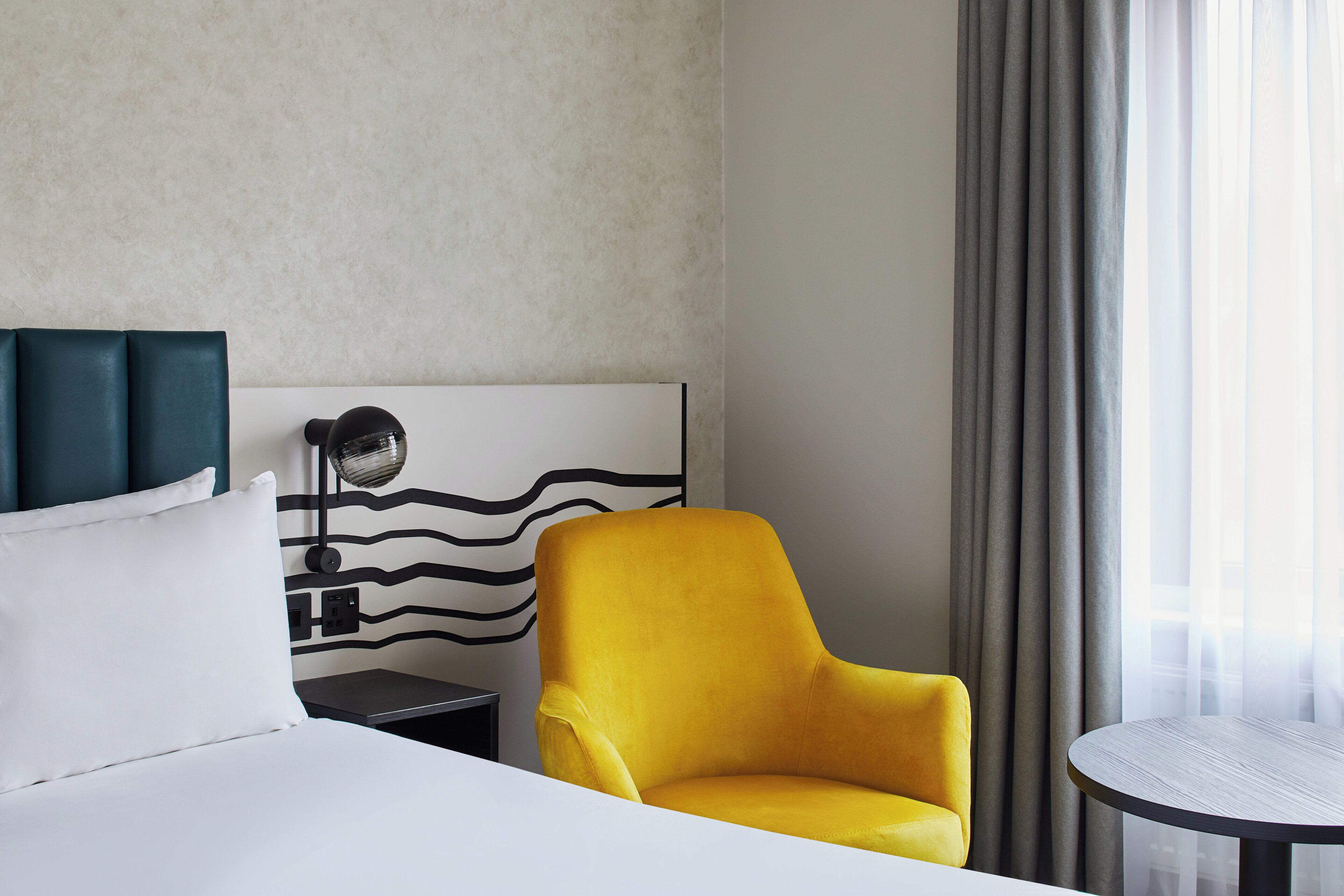 Een hotelkamer met een bed, een gele fauteuil, een ronde tafel en een wandlamp.
