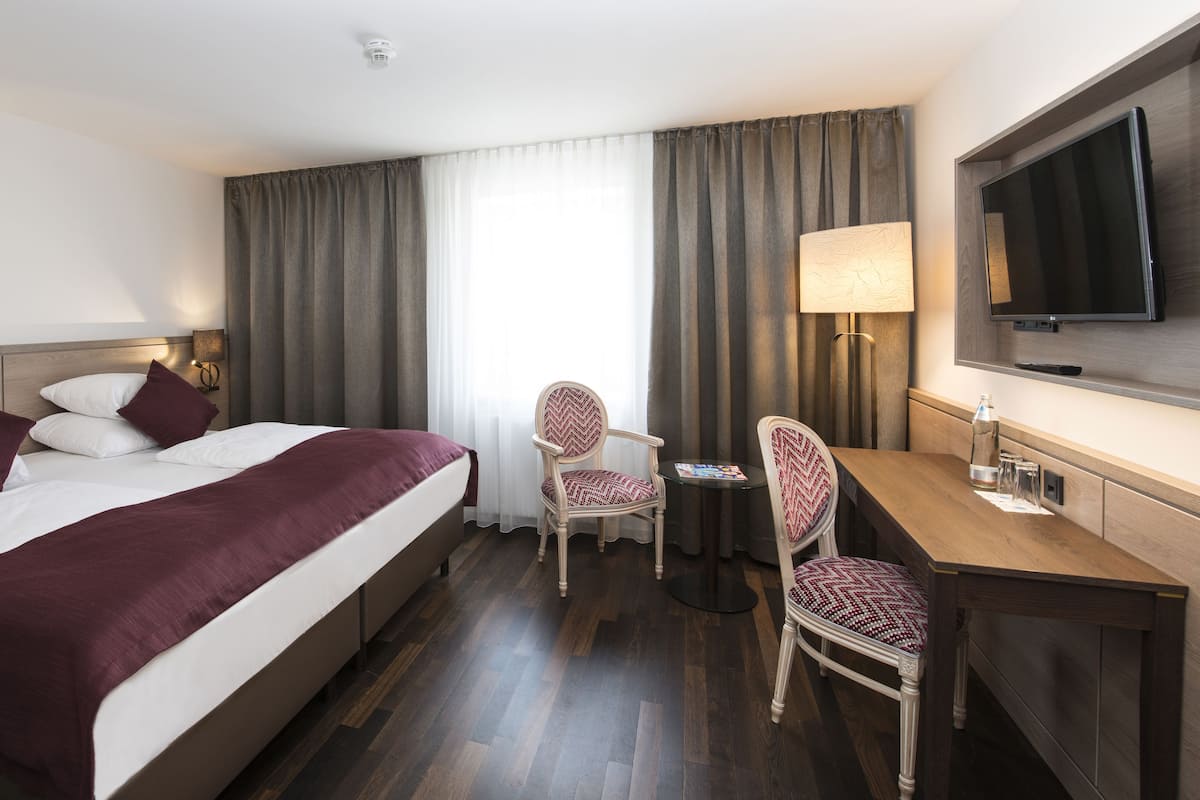 A hotel room with a large bed, a desk, two chairs, a TV, and a window with curtains.