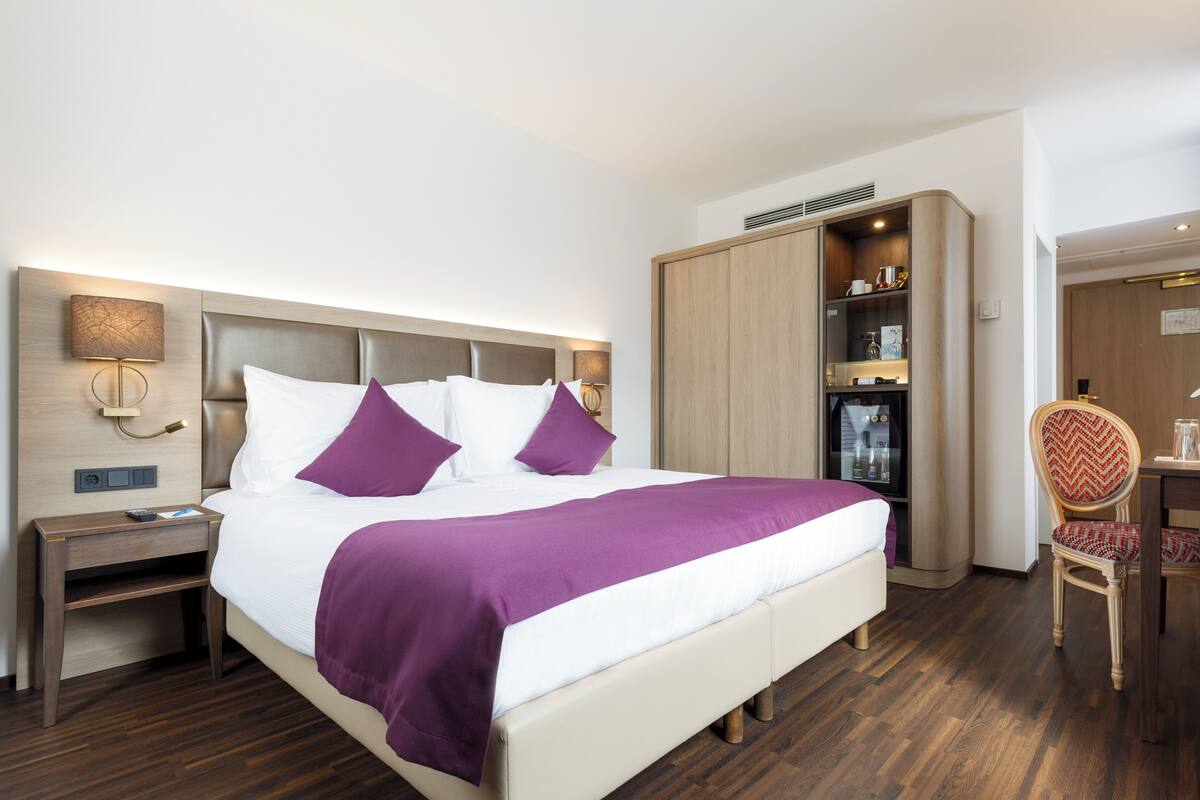 A hotel room with a bed, nightstand, wardrobe, desk, chair, and a television.