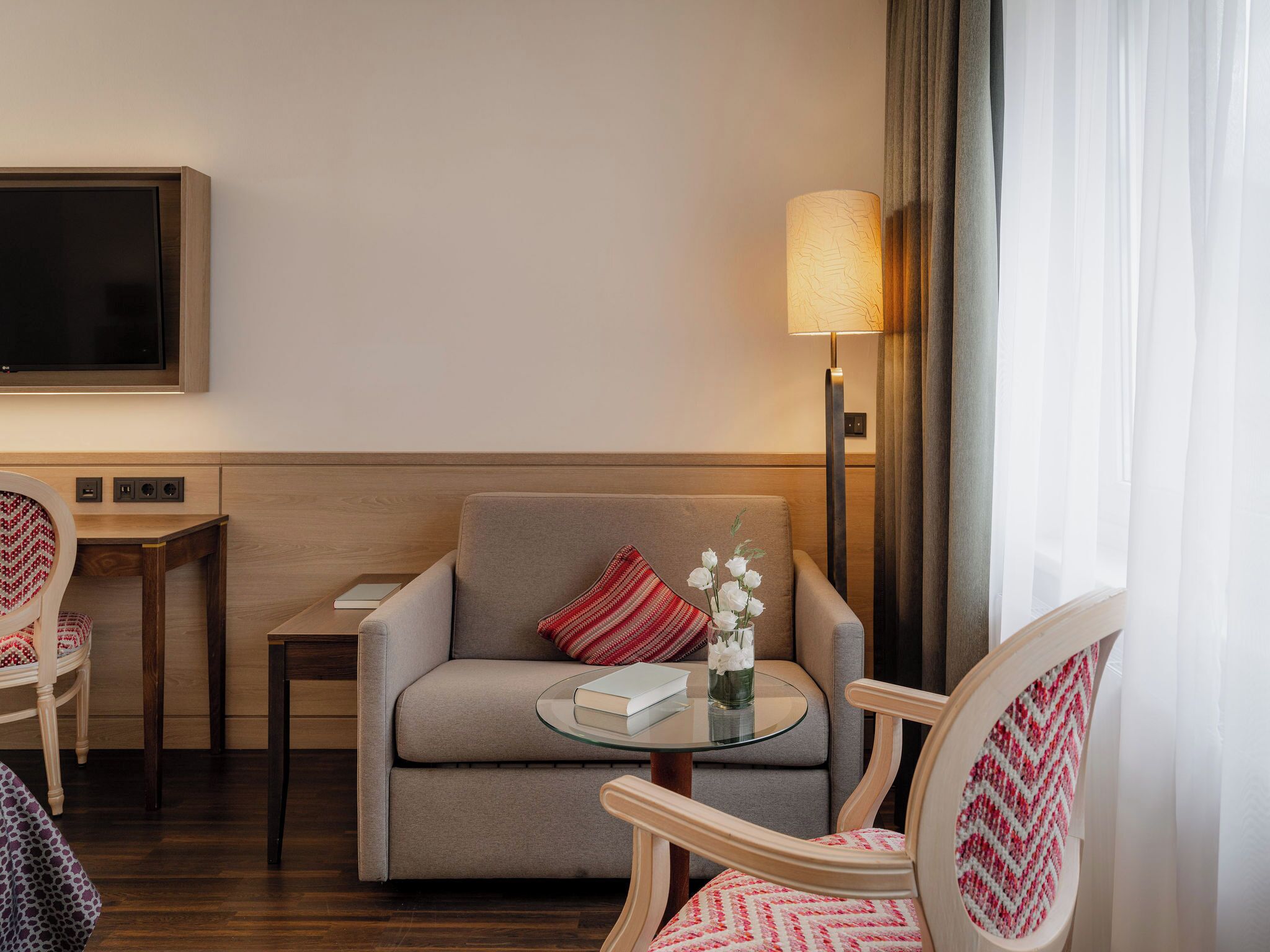 A hotel room with a sofa, a small table with a book, a lamp, and a TV mounted on the wall.
