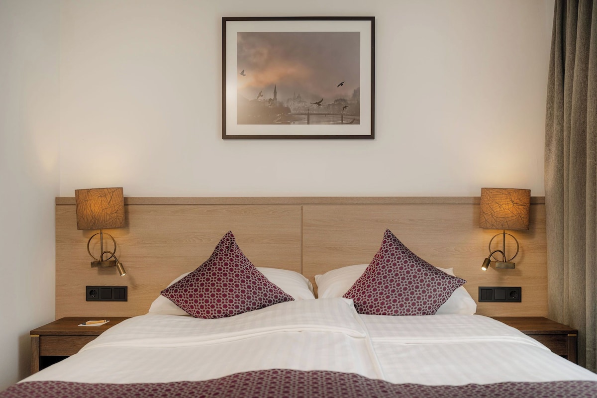 Une chambre d’hôtel avec un grand lit, deux lampes de chevet, un tableau encadré au mur et une fenêtre avec des rideaux.