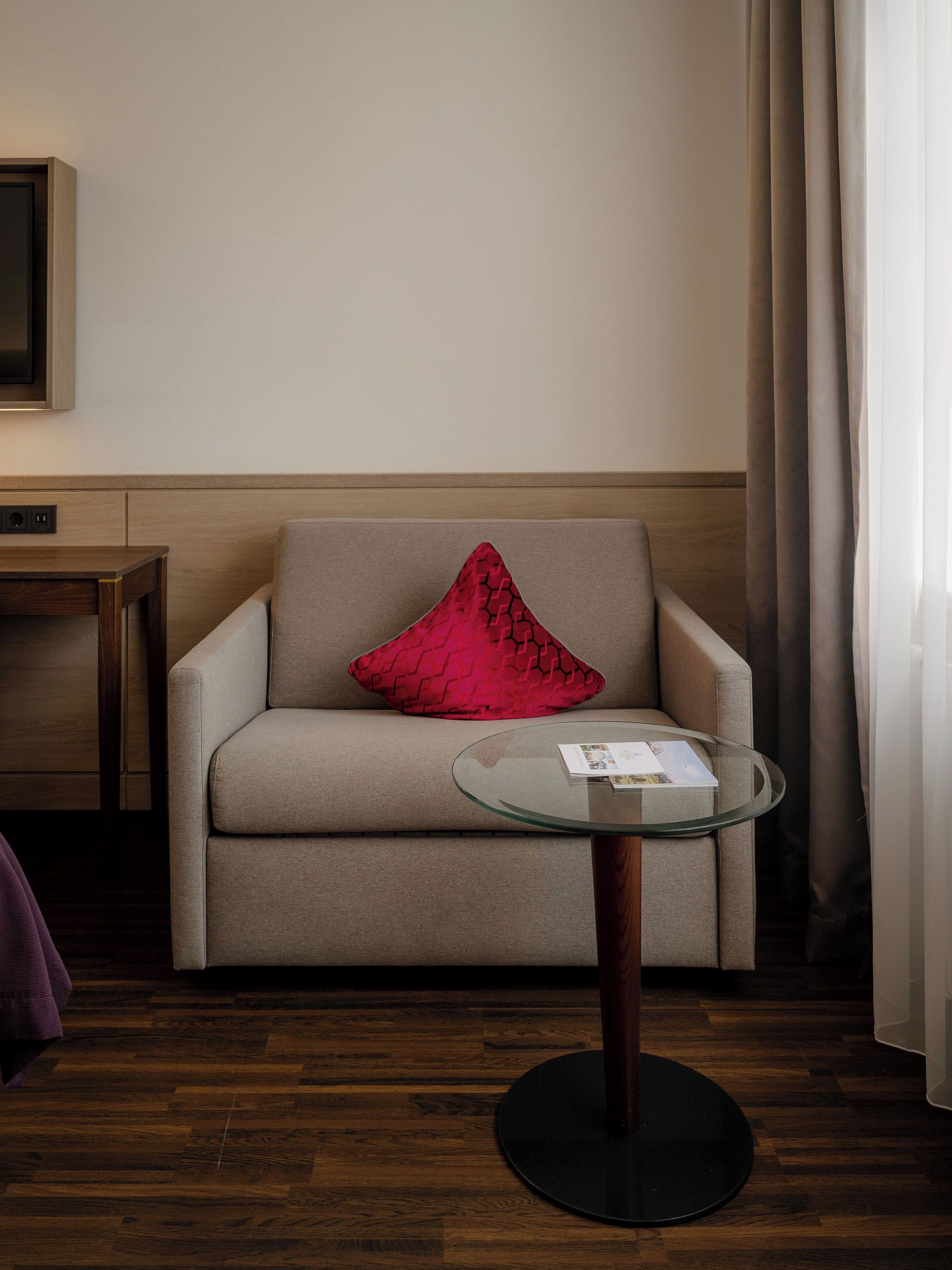 A hotel room with a sofa, a glass coffee table, a red pillow, a wooden floor, and a wall-mounted TV.
