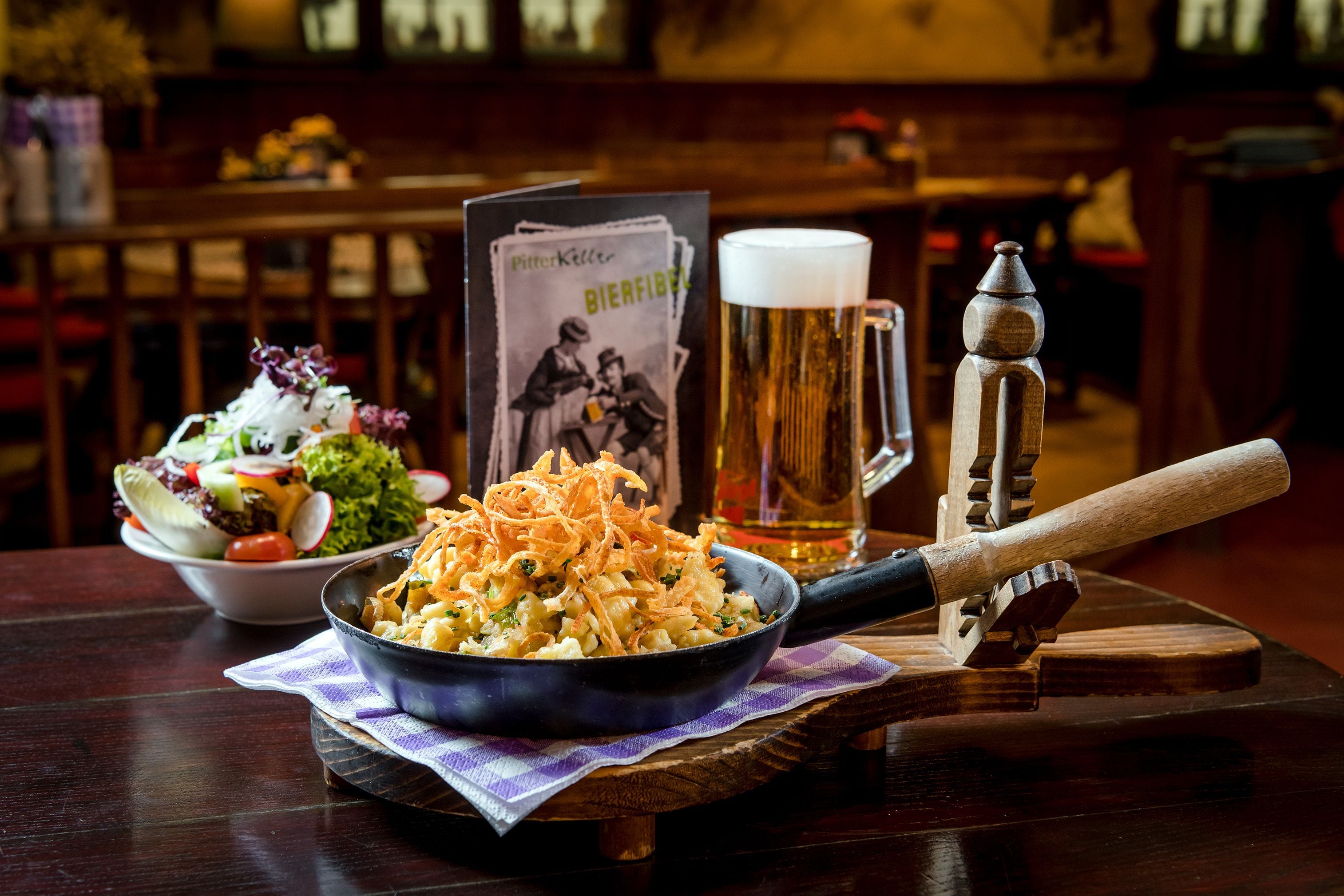 A wooden table with a bowl of salad, a pan of food, a beer mug, and a menu.