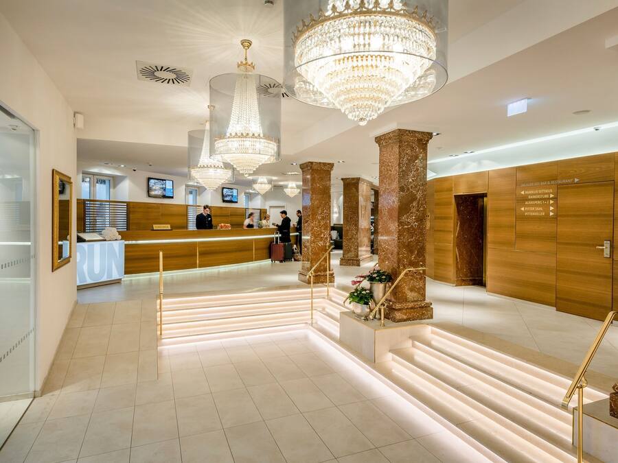 A hotel lobby with a reception desk, large chandeliers, marble columns, and a staircase with illuminated steps.