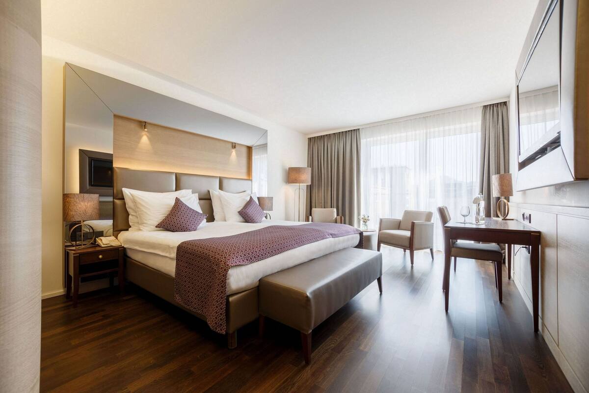 A modern hotel room with a large bed, a desk, a chair, and a bench.