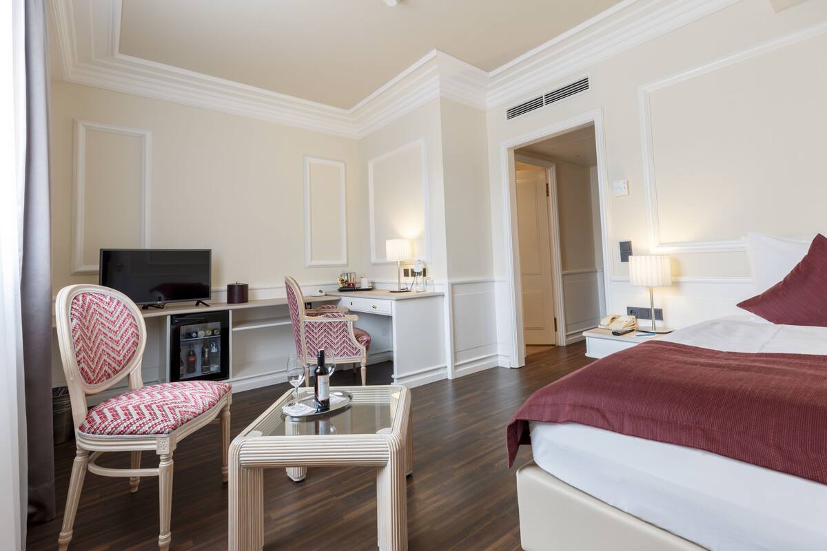 A hotel room with a bed, a desk with a chair, a television, a minibar, and a lamp.