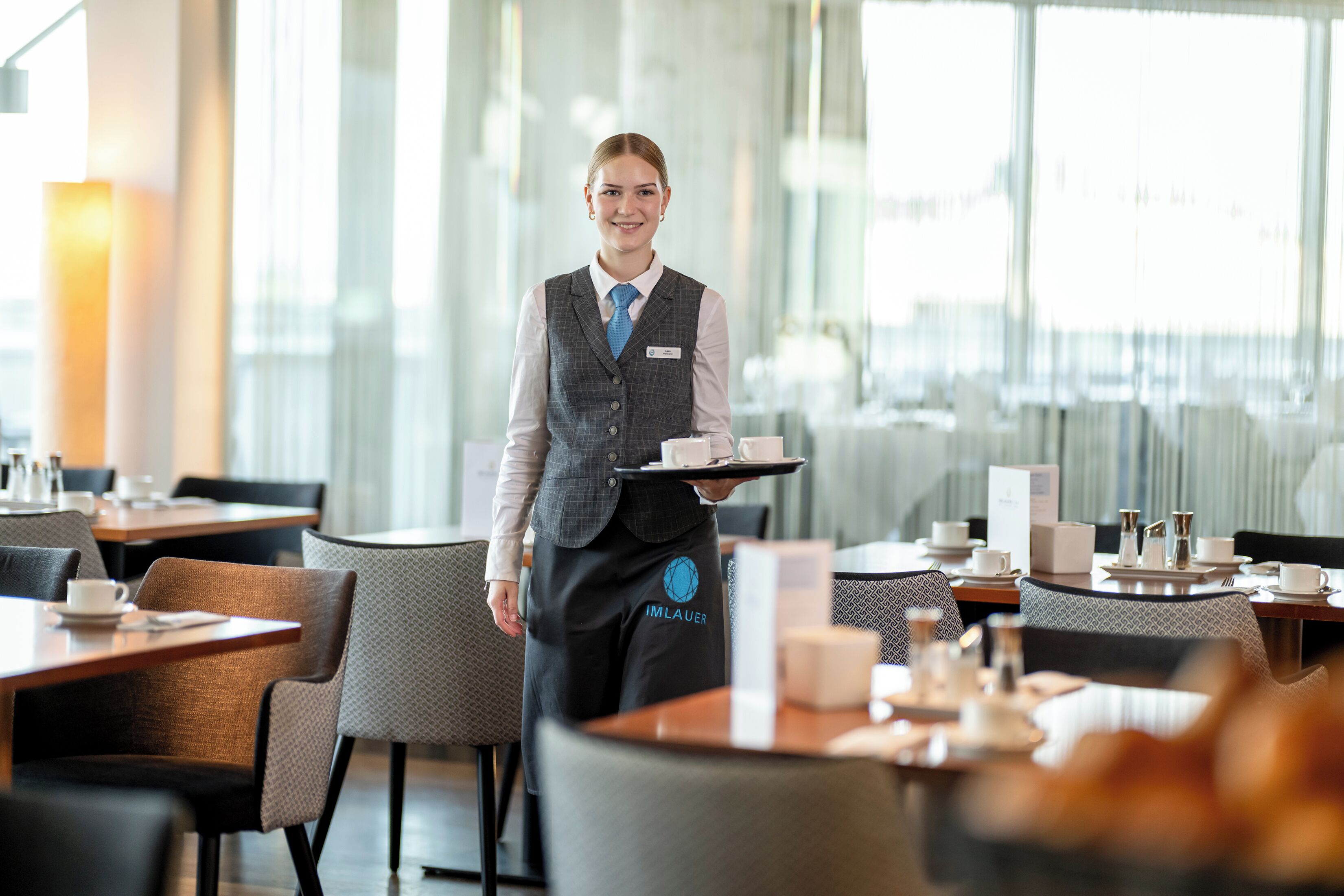 En hotellrestaurang med dukade bord, en servitör i uniform och utsikt över staden genom stora fönster.