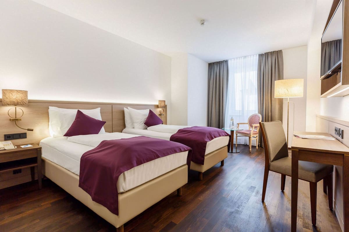 A hotel room with two beds, a desk, a chair, and a window with curtains.