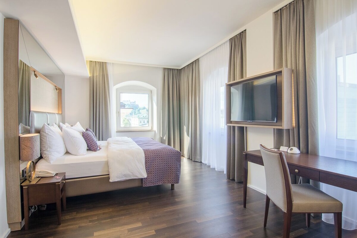 A hotel room with a bed, a desk, a TV, and a window with curtains.