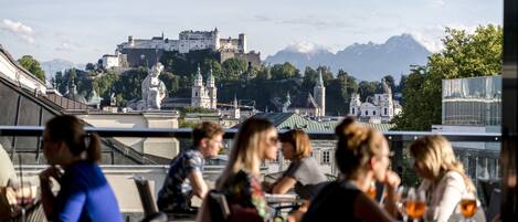 A restaurant with outdoor seating overlooking a historic castle and mountains.