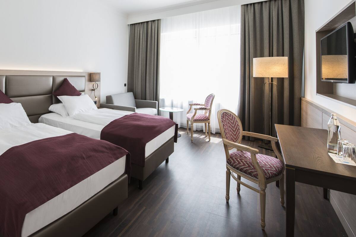 A hotel room with two beds, a desk, a chair, and a TV.