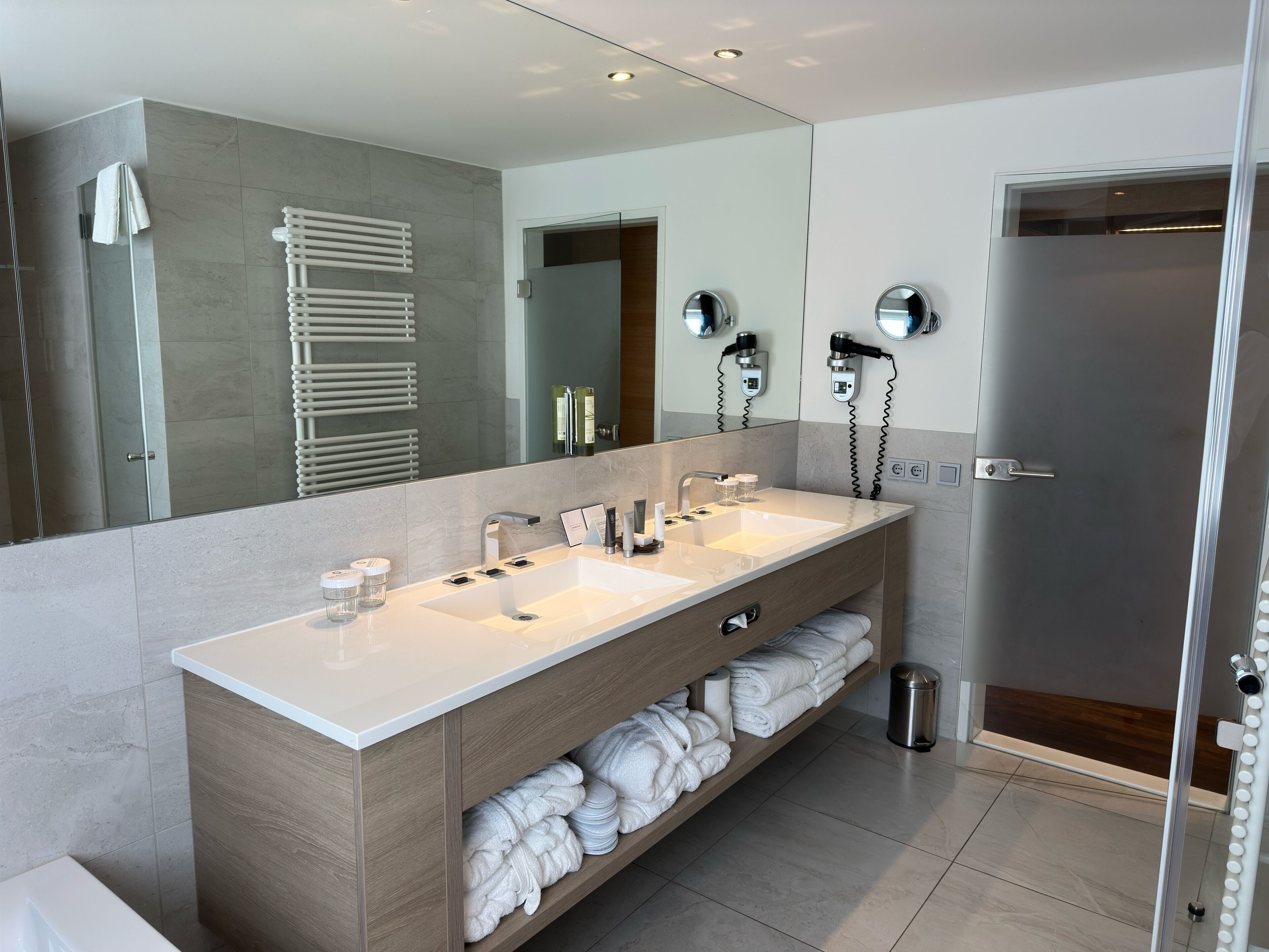 A modern bathroom with a large mirror, two sinks, a towel rack, and a shower area.