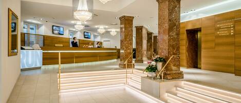 A hotel reception area with a wooden reception desk, marble columns, and a staircase with illuminated steps.