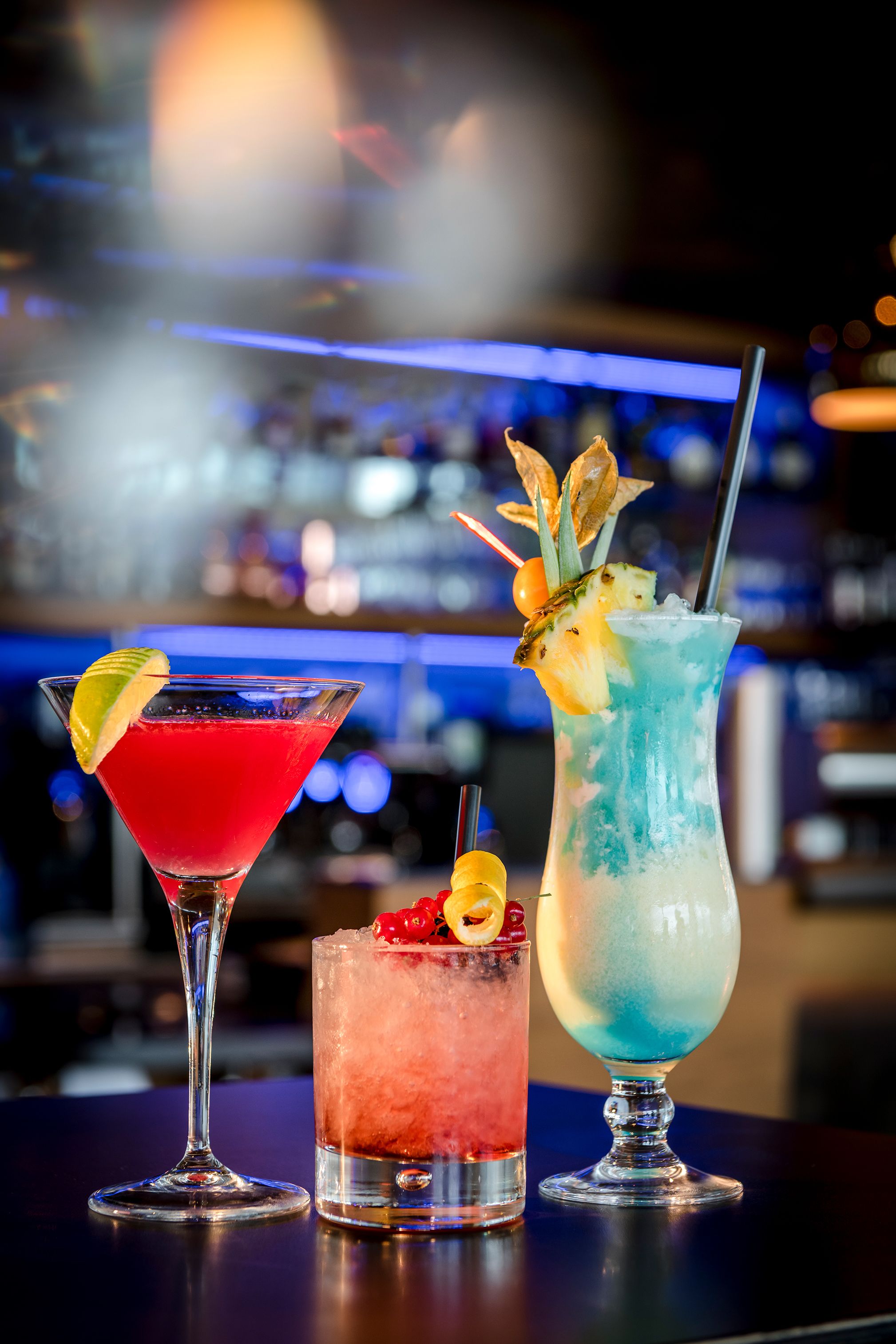 Three cocktails on a bar counter with a blurred background of shelves and bottles.