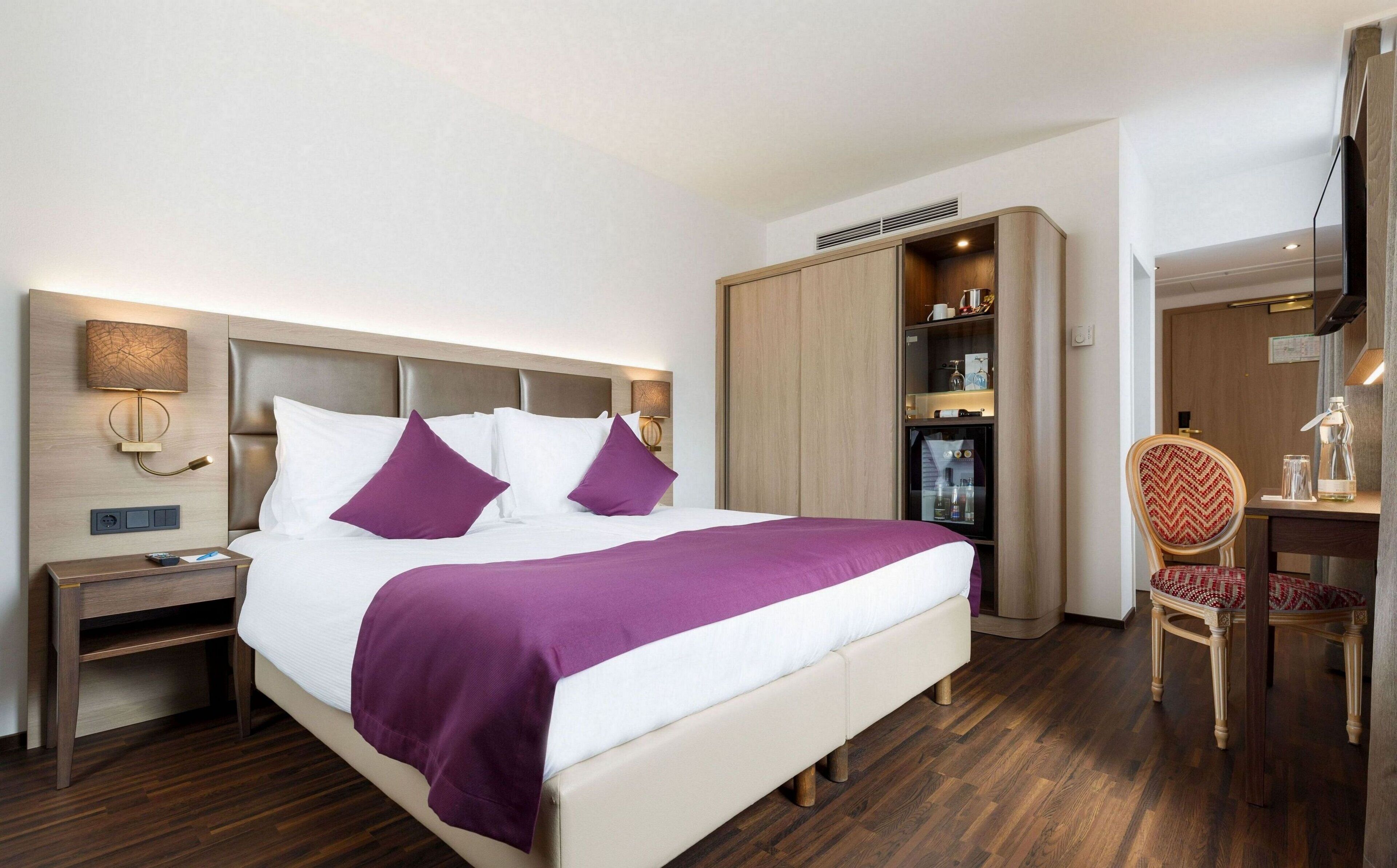 A hotel room with a bed, nightstand, wardrobe, desk, chair, and a television.