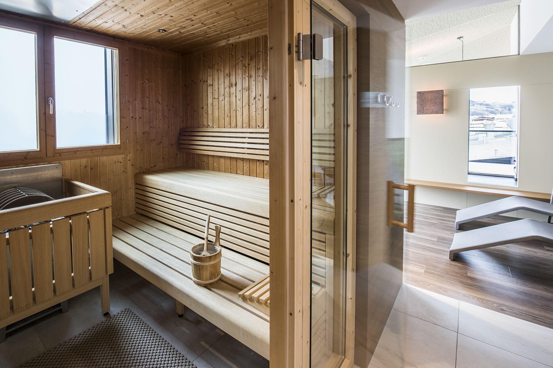 Ein Saunaraum mit Holzbänken, eine Holzkbank mit Eimer und eine Glastür zu einem modernen Badezimmer.