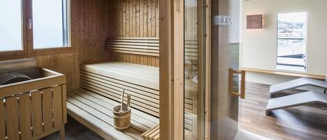 Un sauna con bancos de madera, un banco con un balde y una puerta de cristal que da a un baño moderno.