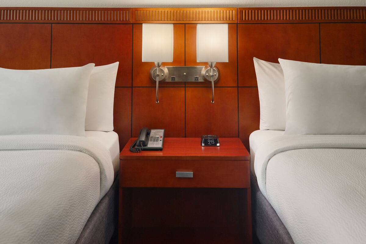 A hotel room with two beds, a nightstand with a phone and a clock, and wall-mounted lamps.