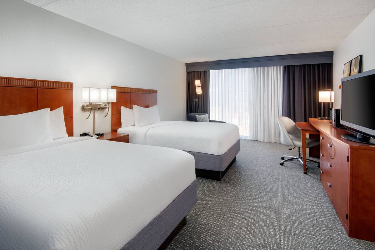 A hotel room with two beds, a desk with a TV, a chair, and a large window with curtains.