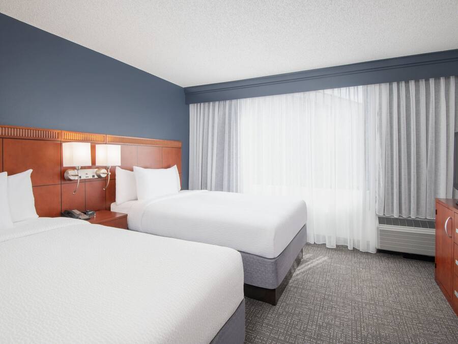 A hotel room with two beds, a wooden headboard, a flat-screen TV, a nightstand, and a large window with sheer curtains.
